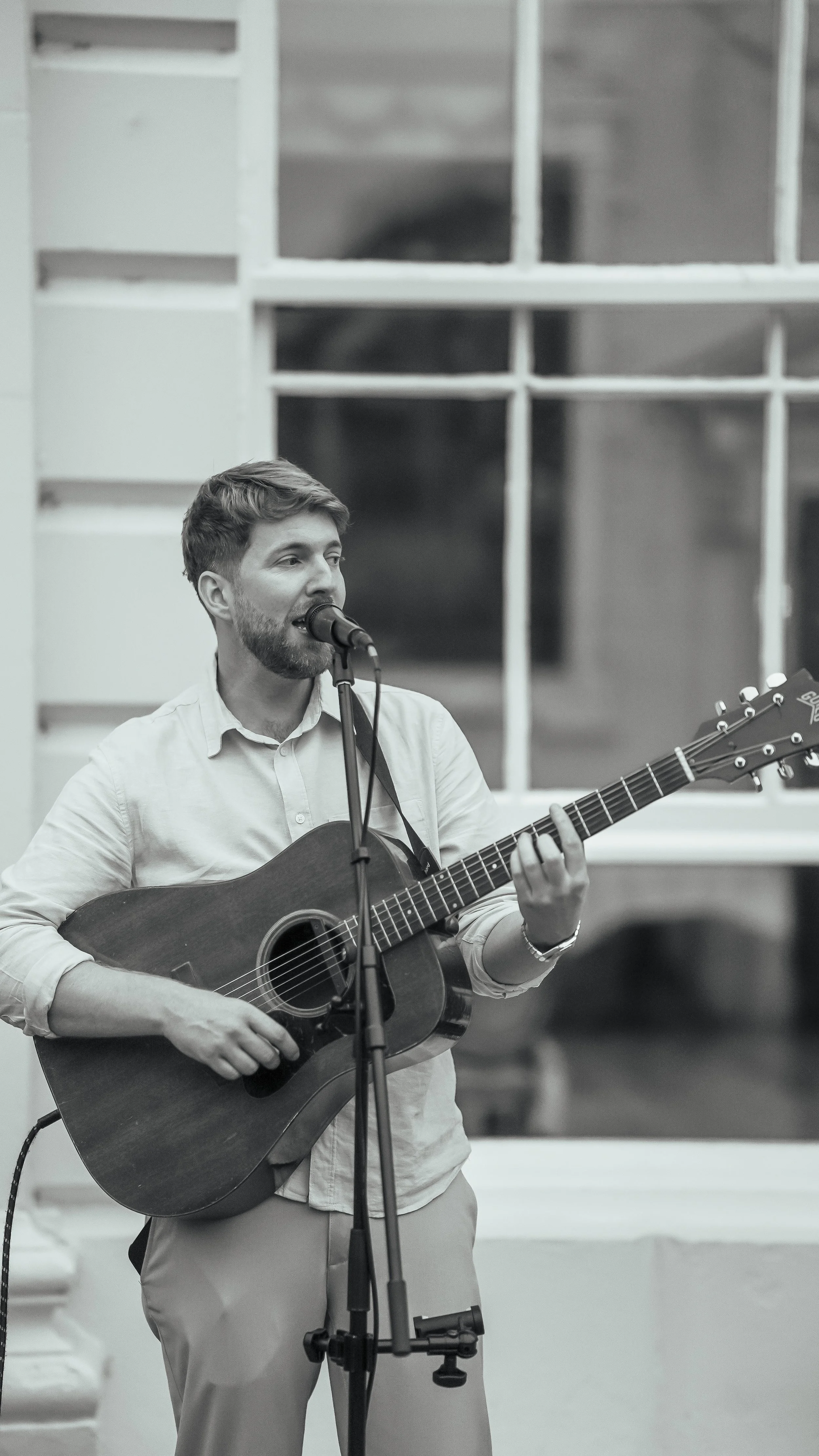 A young man with short hair and a beard, dressed in a light-colored shirt and pants, playing an acoustic guitar and singing into a microphone in an indoor setting.