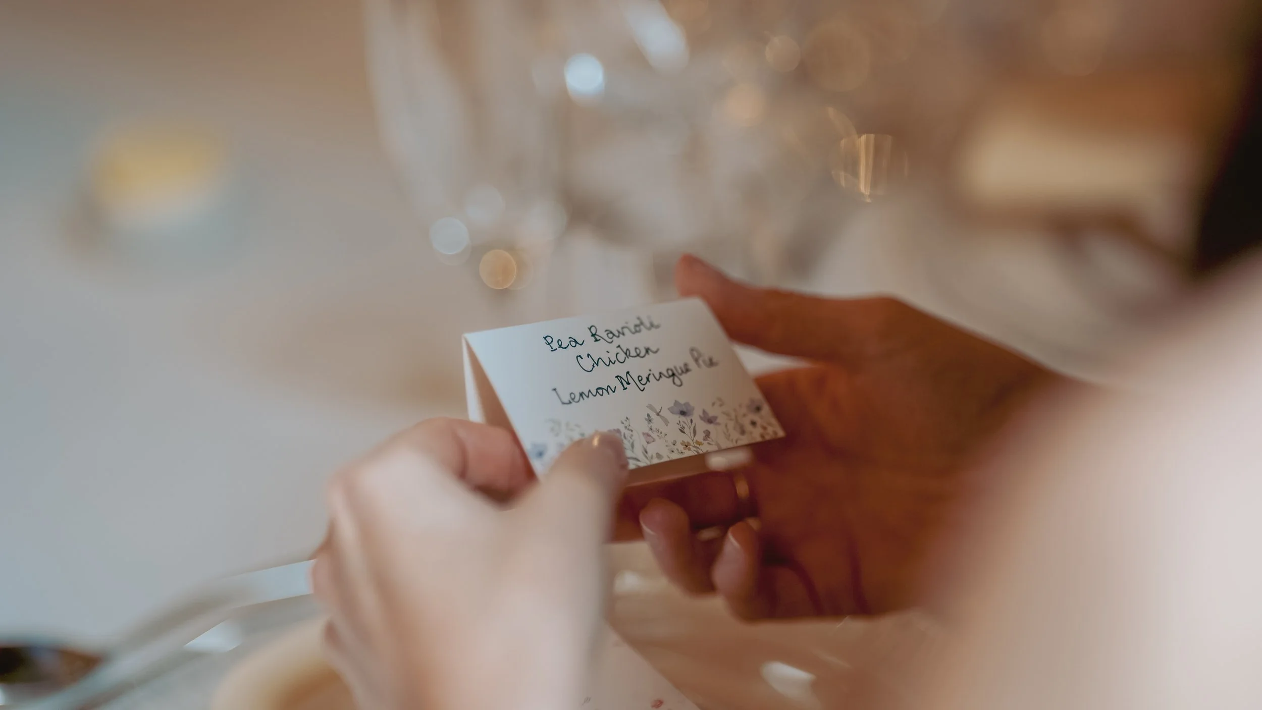 Close-up of a person holding a small white card with handwritten menu items, including pea ravioli, chicken, lemon meringue pie, on a table with glasses and blurred background.