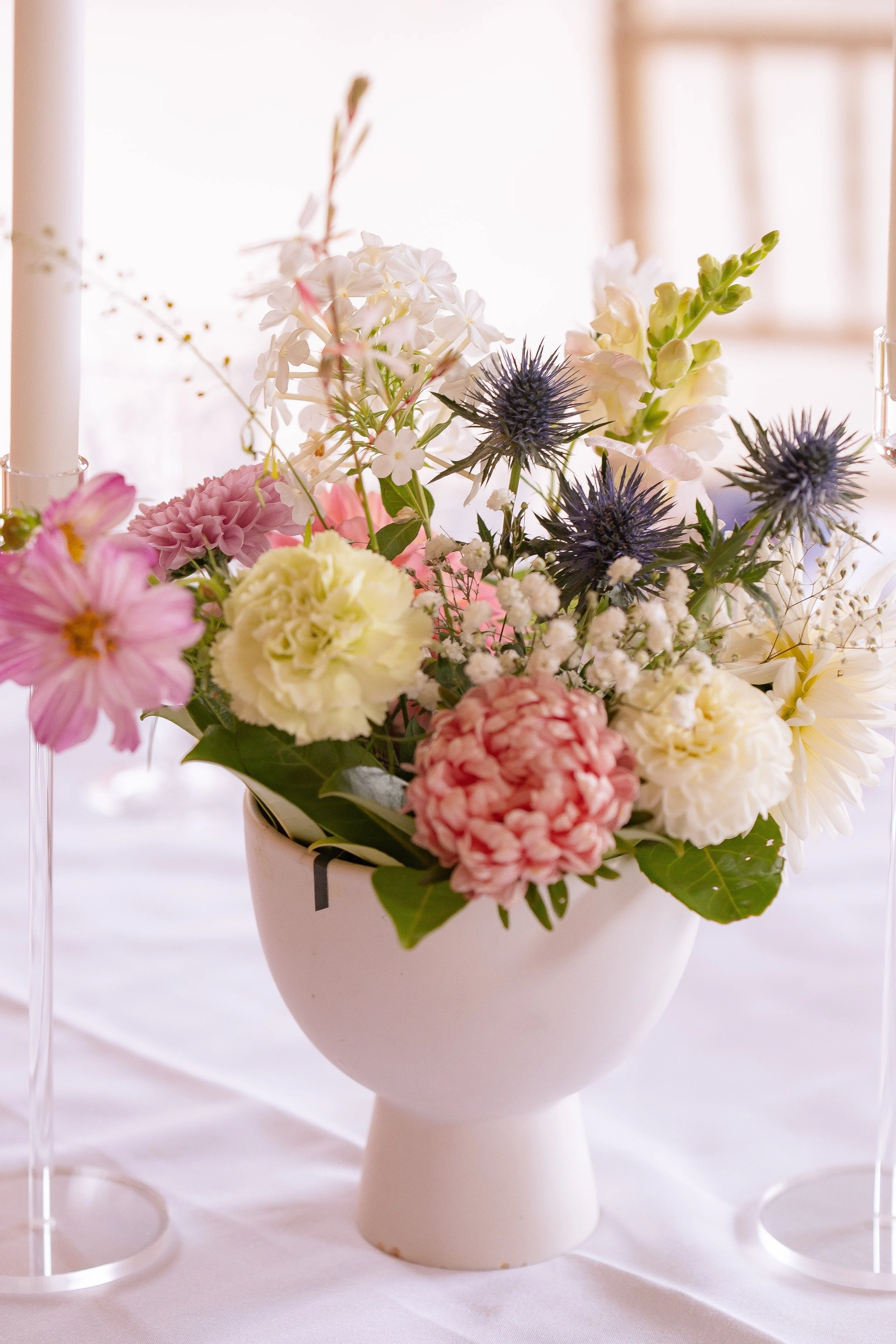 A white ceramic vase filled with a colorful arrangement of various flowers including pink, white, and purple blooms on a white tablecloth, with candles in glass holders nearby, in a softly lit room.