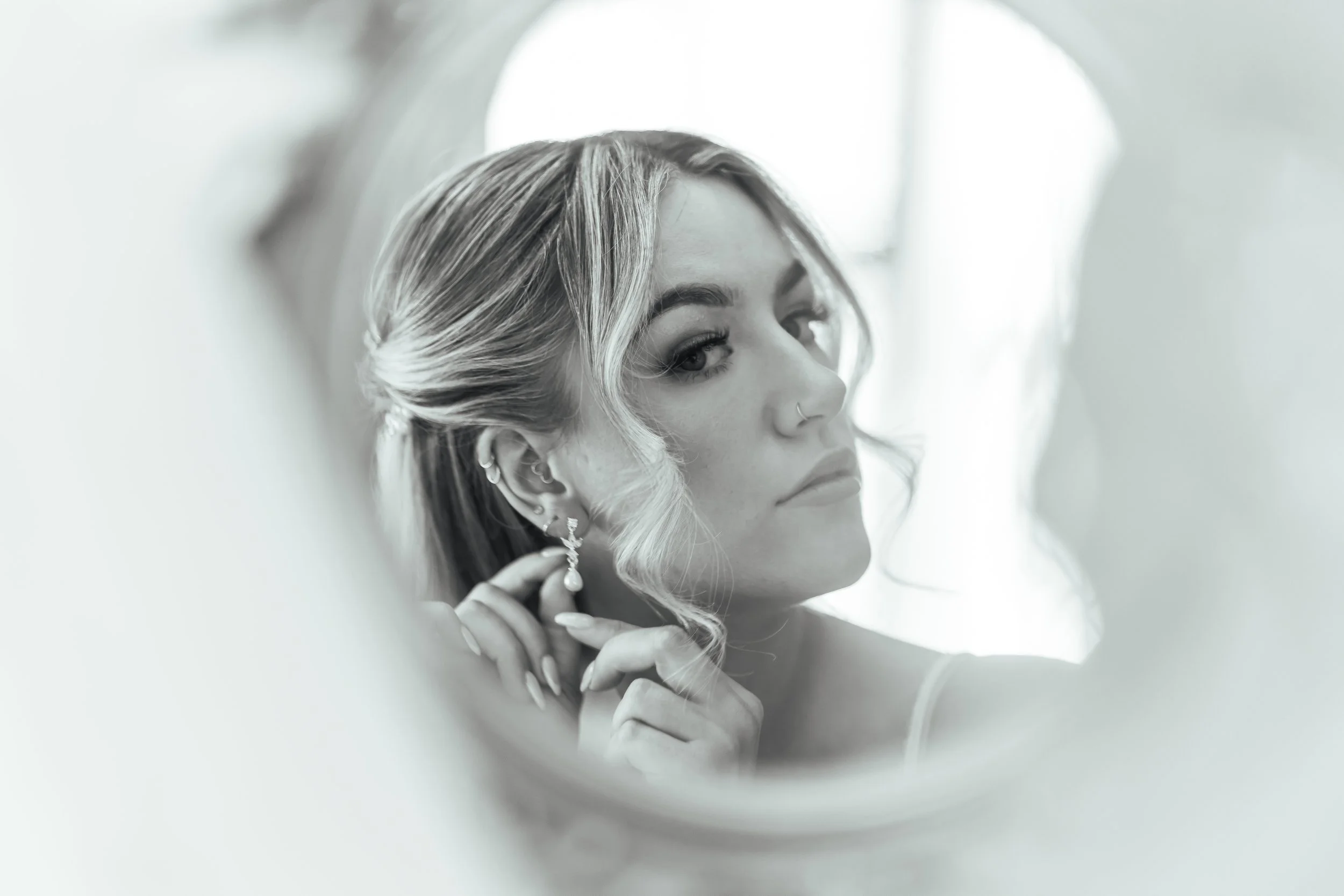 Black-and-white photo of a woman with styled hair and earrings, looking at her reflection in a mirror while adjusting her earring.
