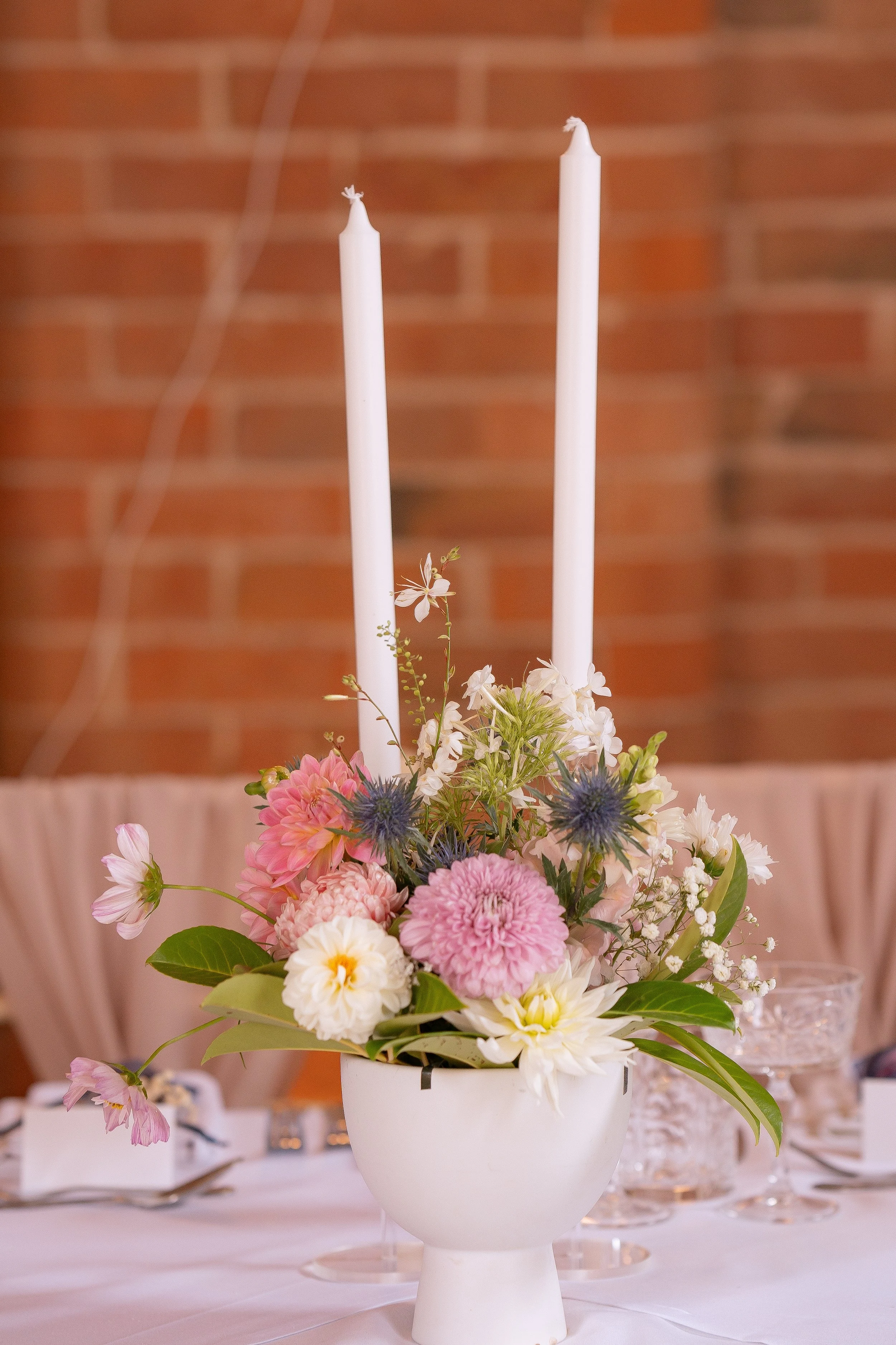 A floral centerpiece with pink, white, and purple flowers and two tall white candles on a table.