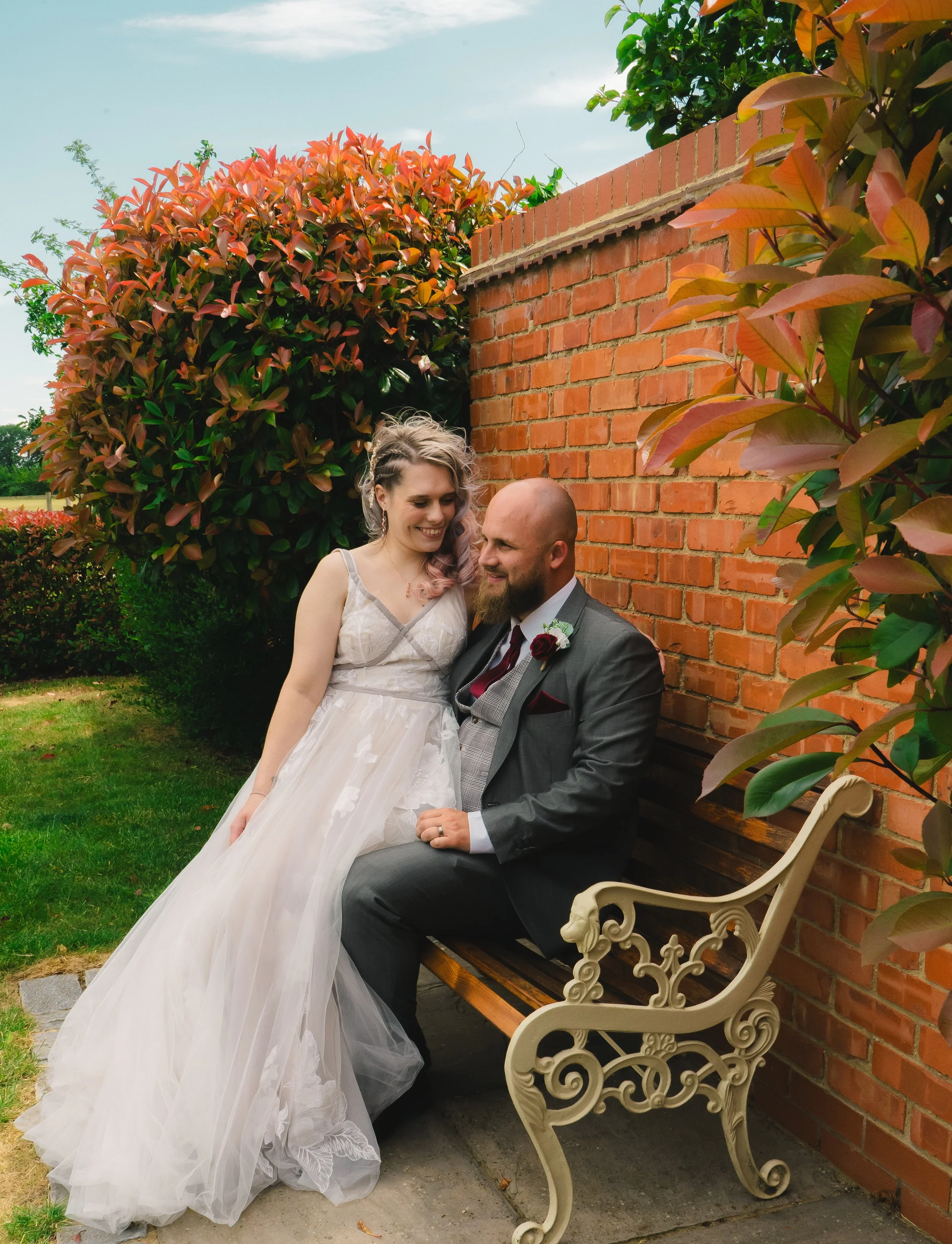 A bride and groom sitting on a bench outside, smiling at each other. The bride is wearing a white wedding dress, and the groom is wearing a gray suit with a boutonniere. Behind them are lush bushes and a brick wall.