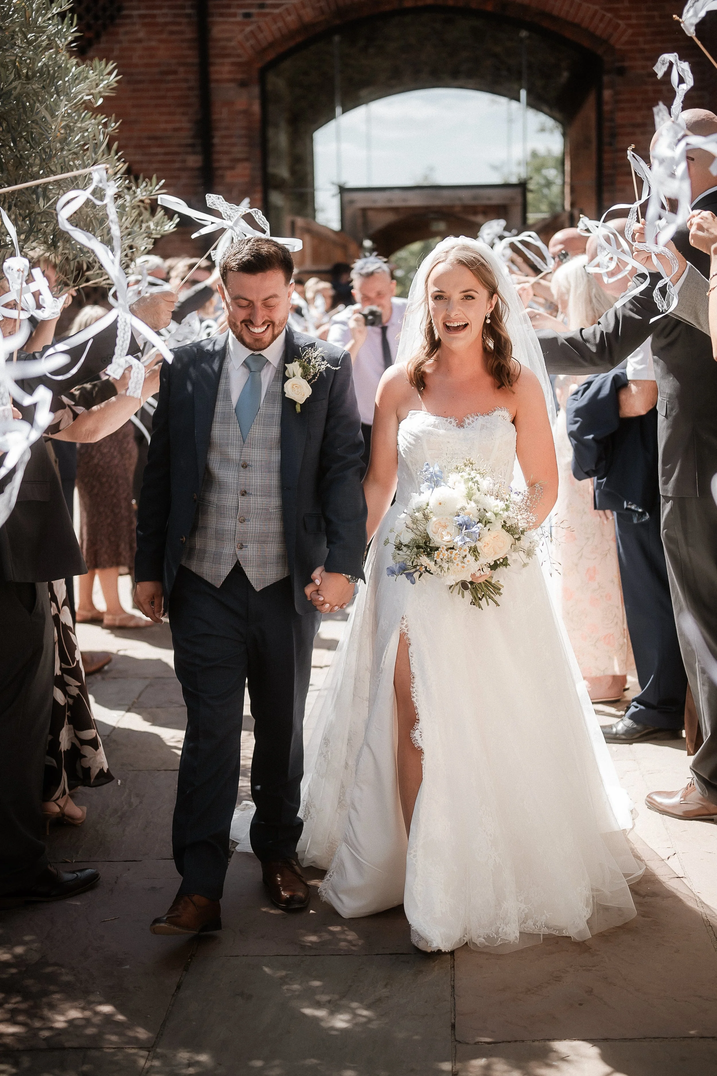 A newlywed couple walking hand in hand after their wedding ceremony, surrounded by guests celebrating and throwing streamers. The bride wears a white lace wedding dress and holding a bouquet, while the groom wears a dark suit with a boutonniere. They