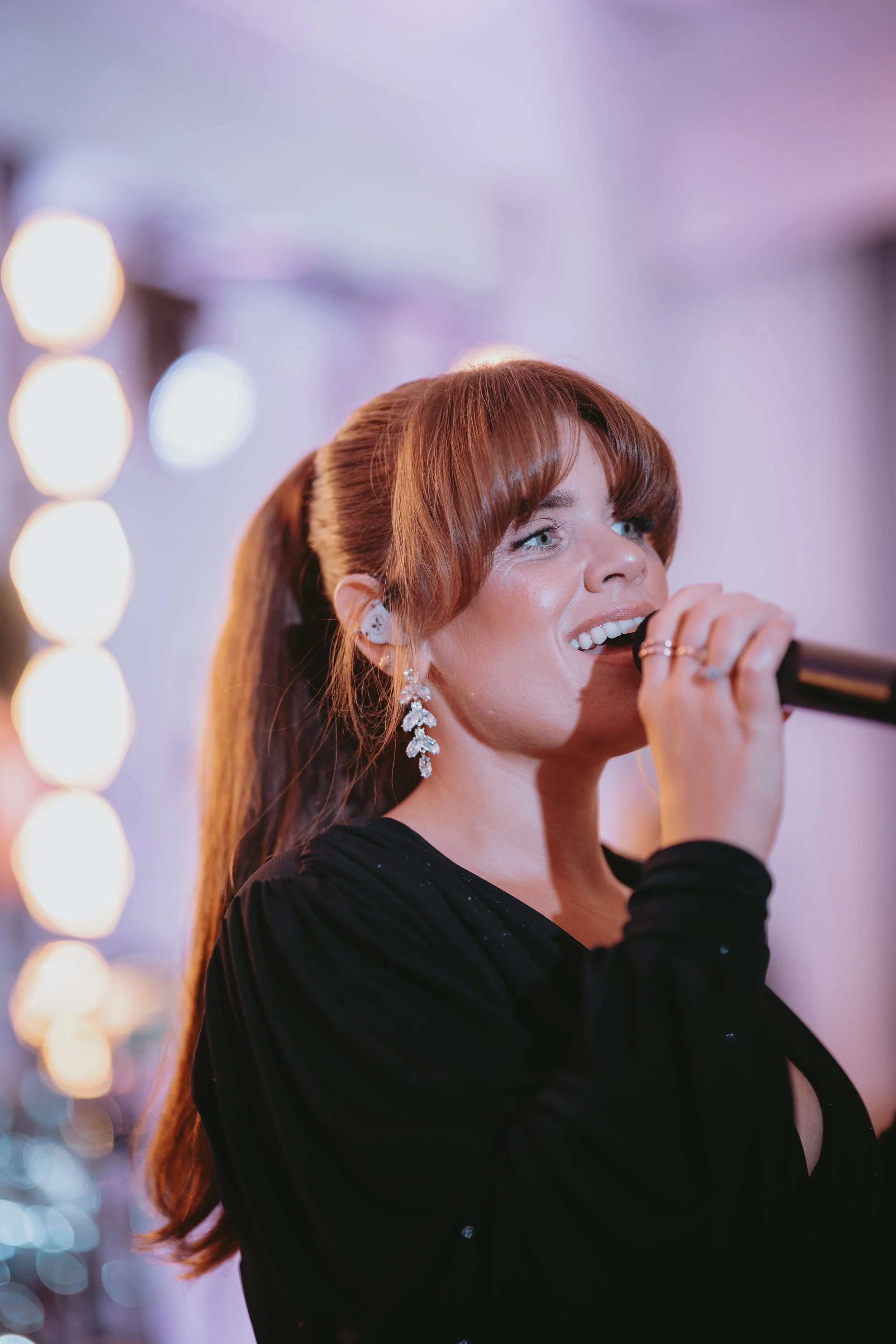 A woman with long, reddish-brown hair singing into a microphone at an event, wearing earrings and a black dress.