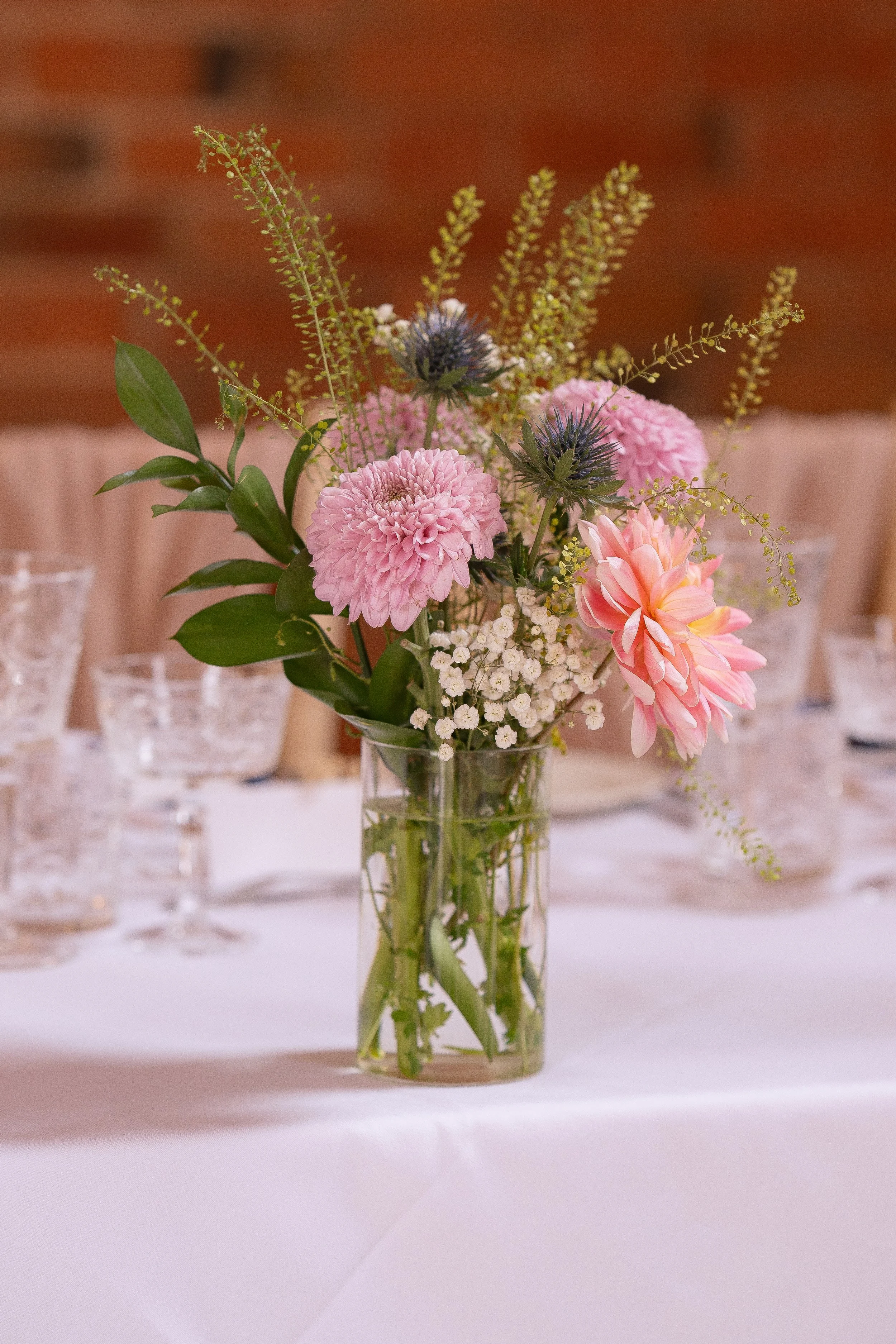 A floral centerpiece with pink, purple, and white flowers in a clear glass vase on a white tablecloth, with glassware and chairs in the background.