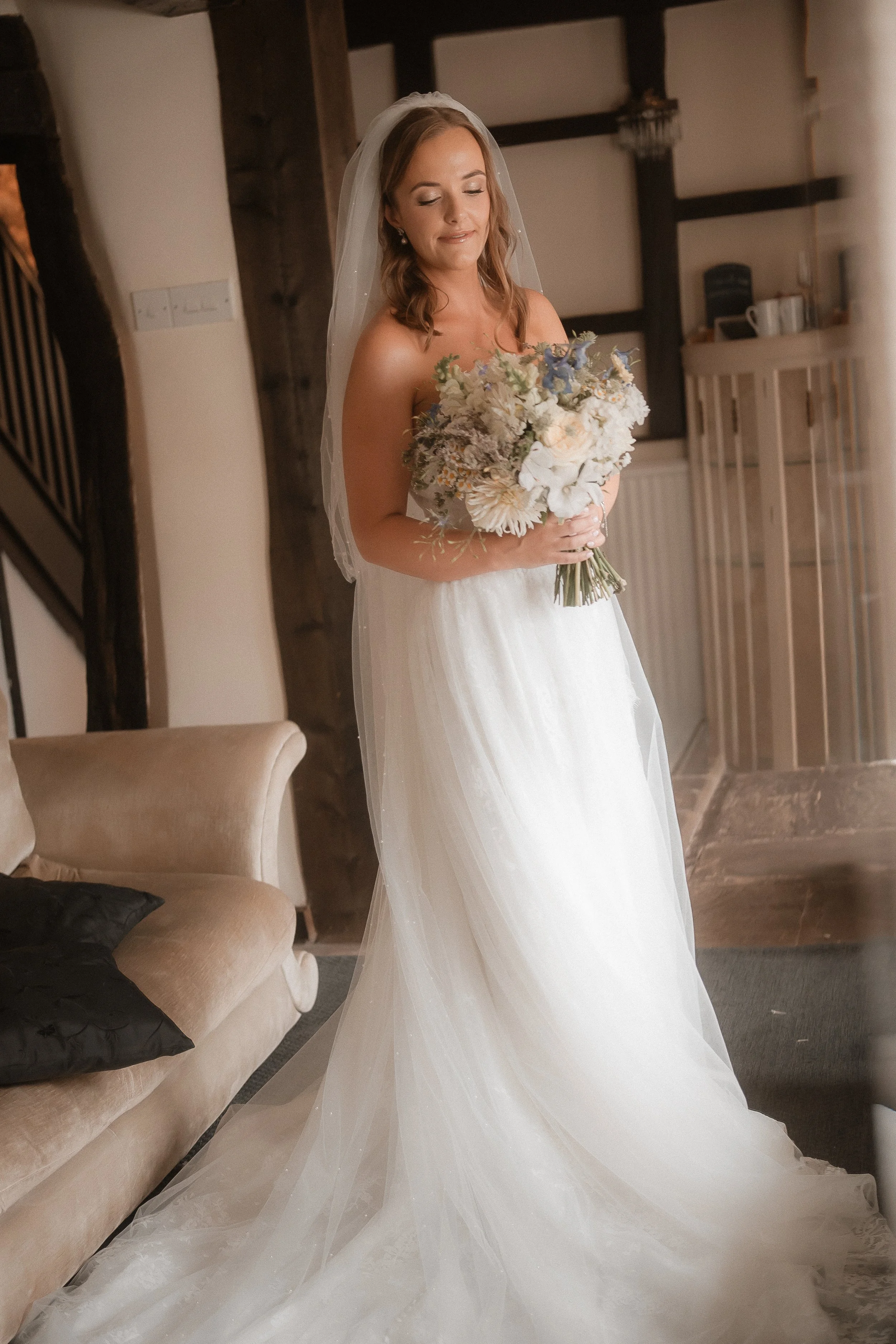 A bride with shoulder-length hair, wearing a veil and a strapless wedding dress, holding a bouquet of white and light purple flowers, standing indoors near a beige sofa with black pillows and a wooden staircase.