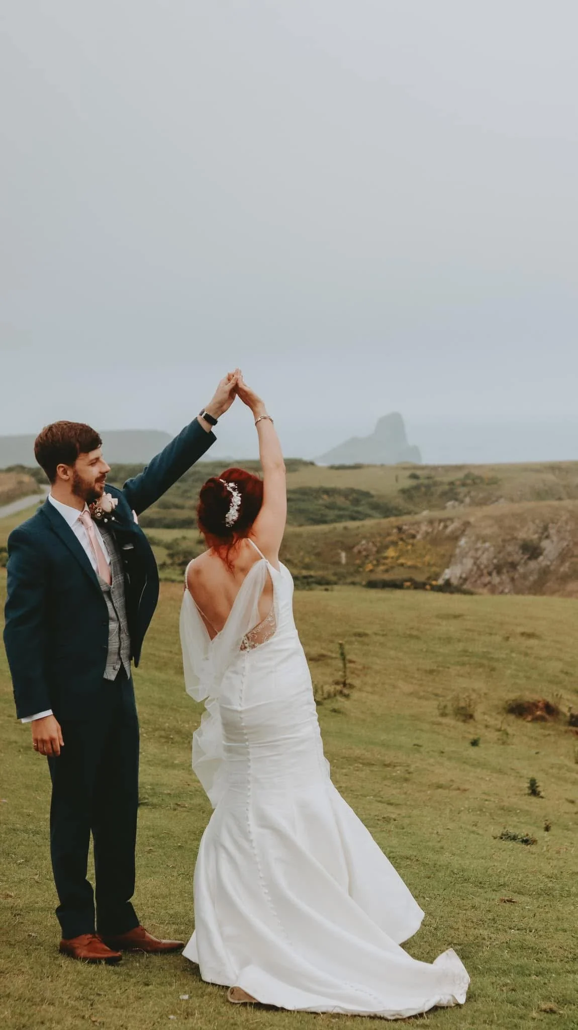 A newlywed couple dancing outdoors, with the groom in a blue suit and the bride in a white wedding dress, set against a scenic grassy landscape.