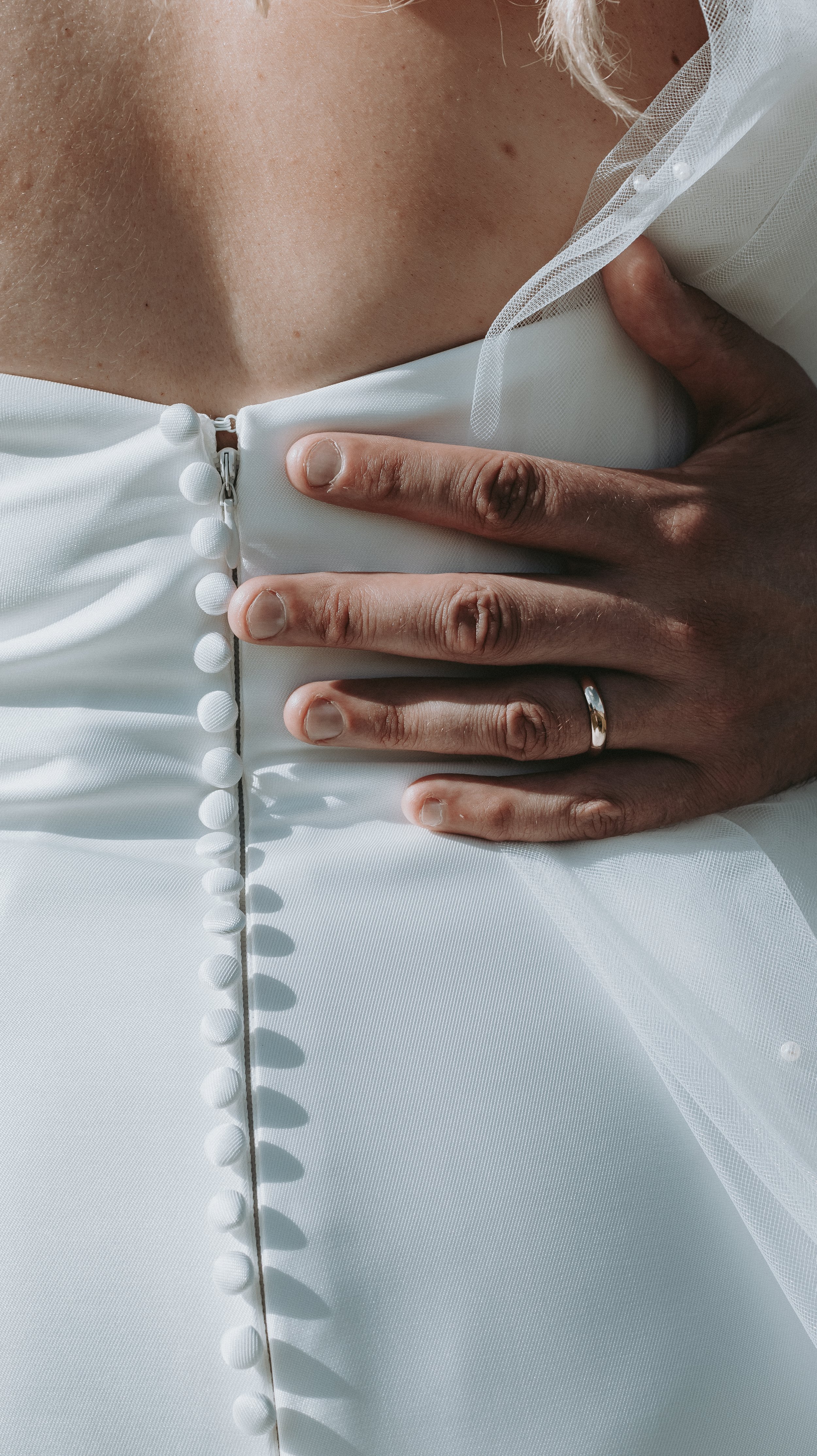 Close-up of a person's hand with a wedding ring resting on the back of a white dress with button details.