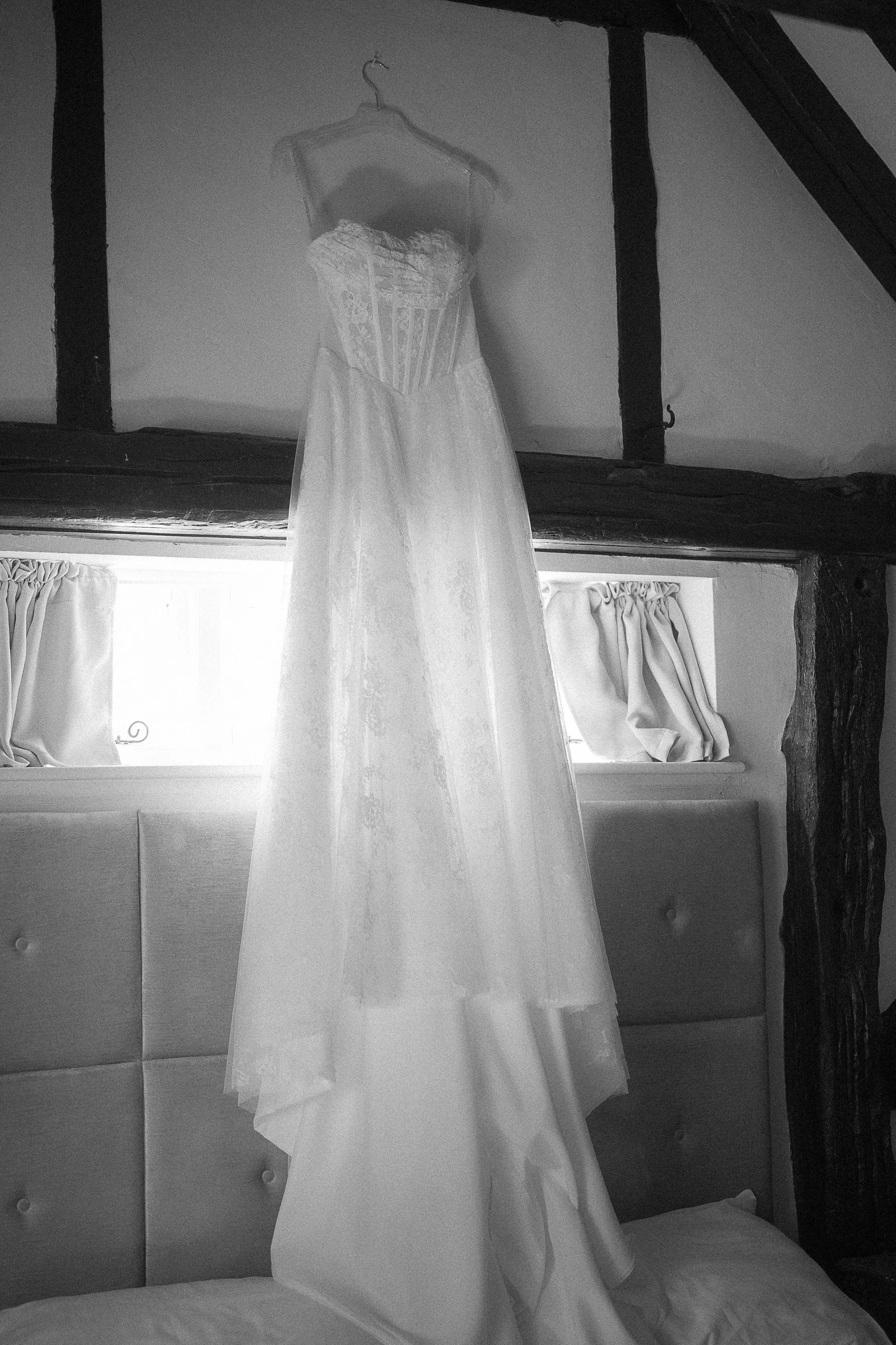 A white wedding dress hanging on a hanger inside a room with wooden beams and curtains.