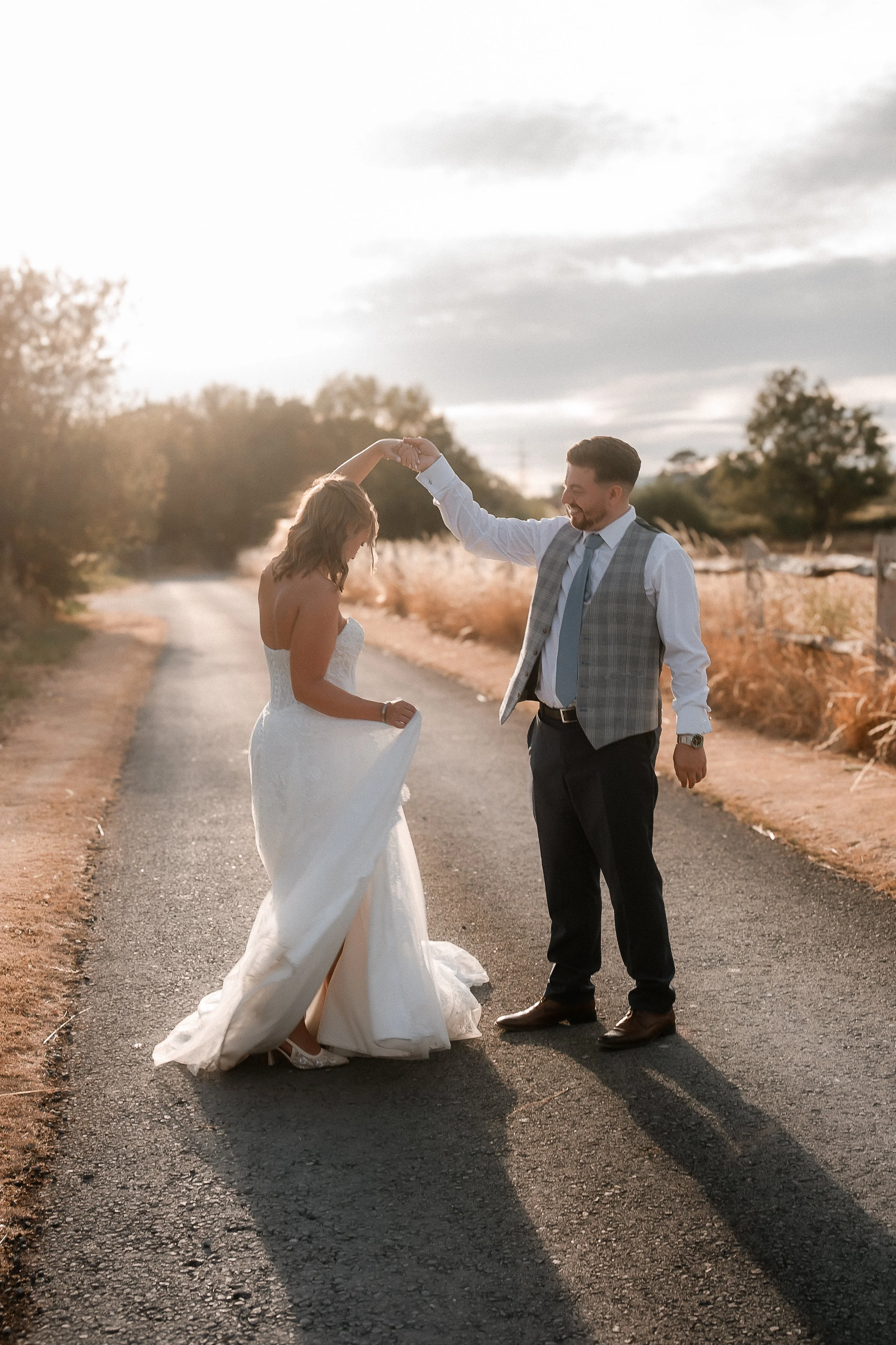 A couple, dressed in wedding attire, dancing on a country road during sunset. The bride is in a white strapless wedding gown, and the groom is in a dress shirt, vest, and tie. The woman is holding up her dress slightly while the man twirls her, smili