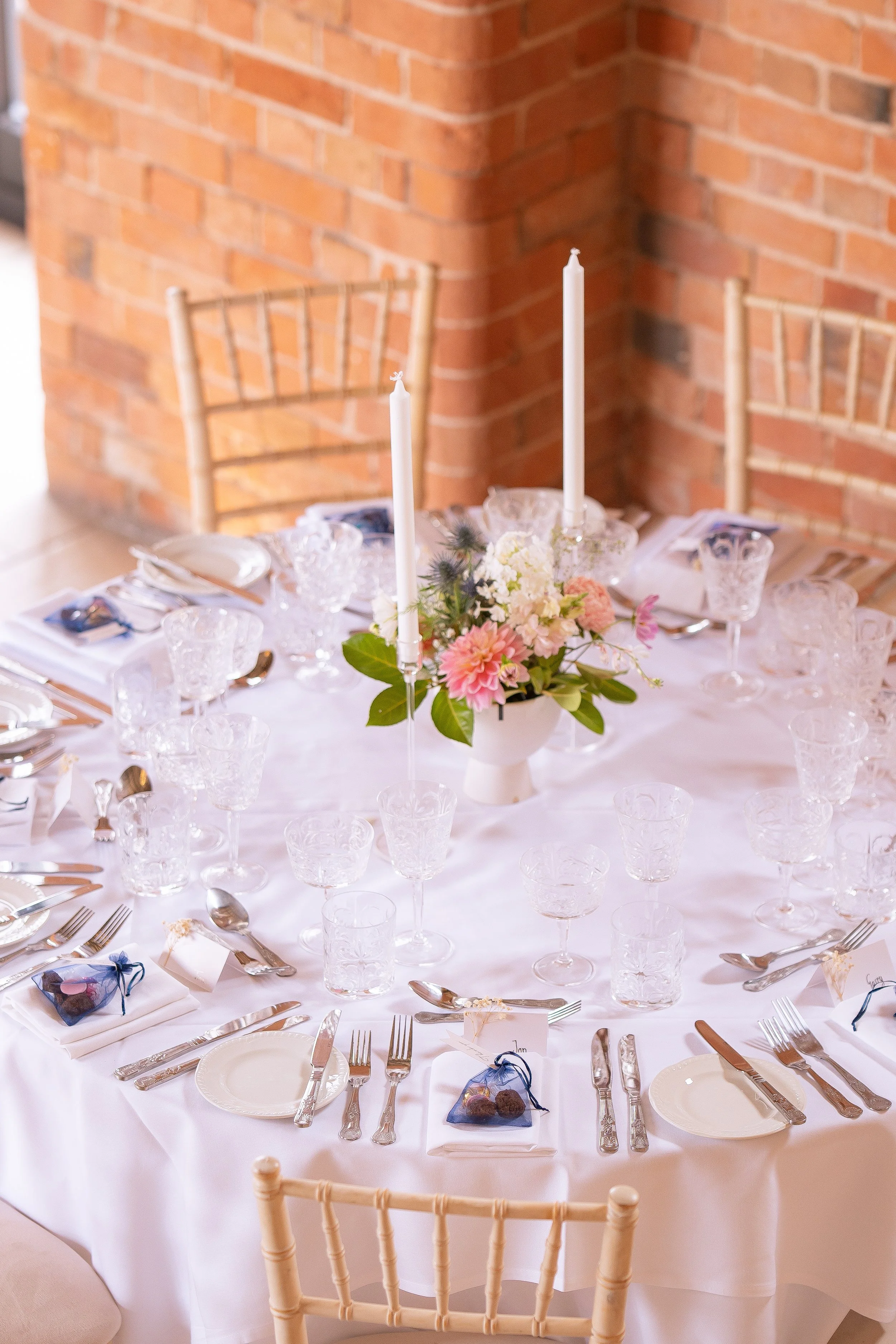 Formal dining table setup with a floral centerpiece, tall candles, glassware, and place settings against a brick wall background.
