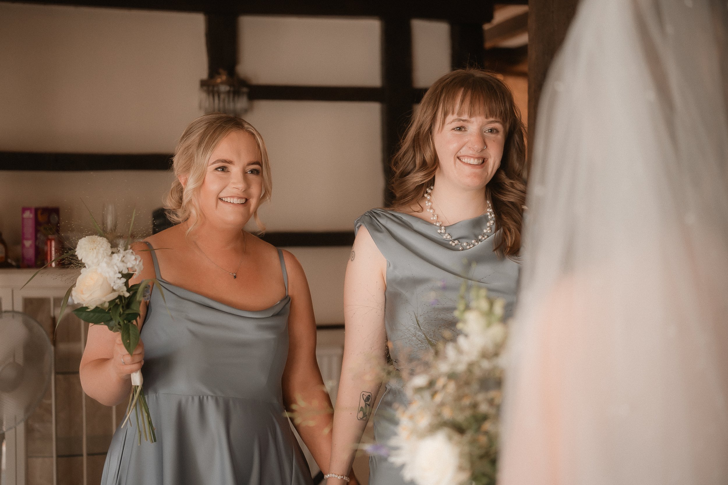 Two women in gray dresses smiling and holding bouquets, standing in a room with wooden beams, during a wedding or special occasion.