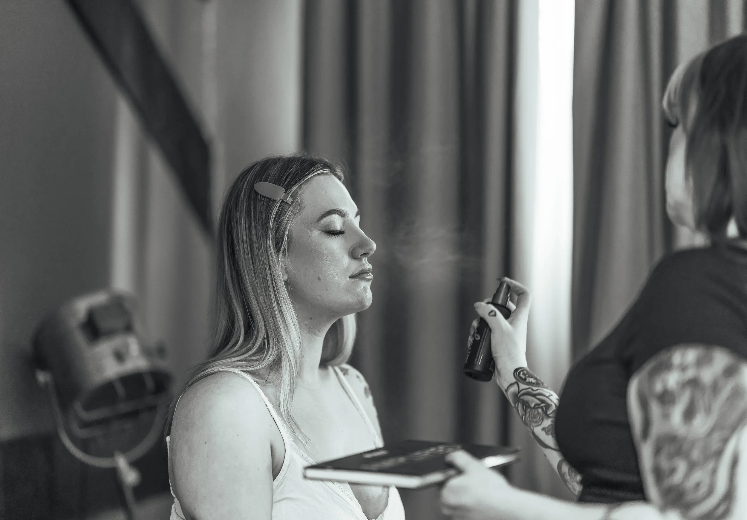 A woman with closed eyes is having makeup or skincare spray applied by a person with tattoos on their arm in a room with curtains.