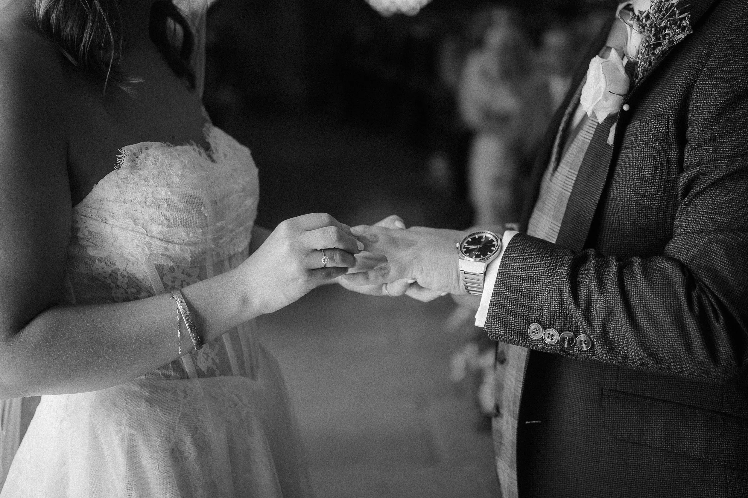 Close-up of a bride and groom holding hands during their wedding ceremony, with the bride wearing a lace wedding dress and the groom in a checkered suit, both wearing watches, and the bride's engagement ring visible.