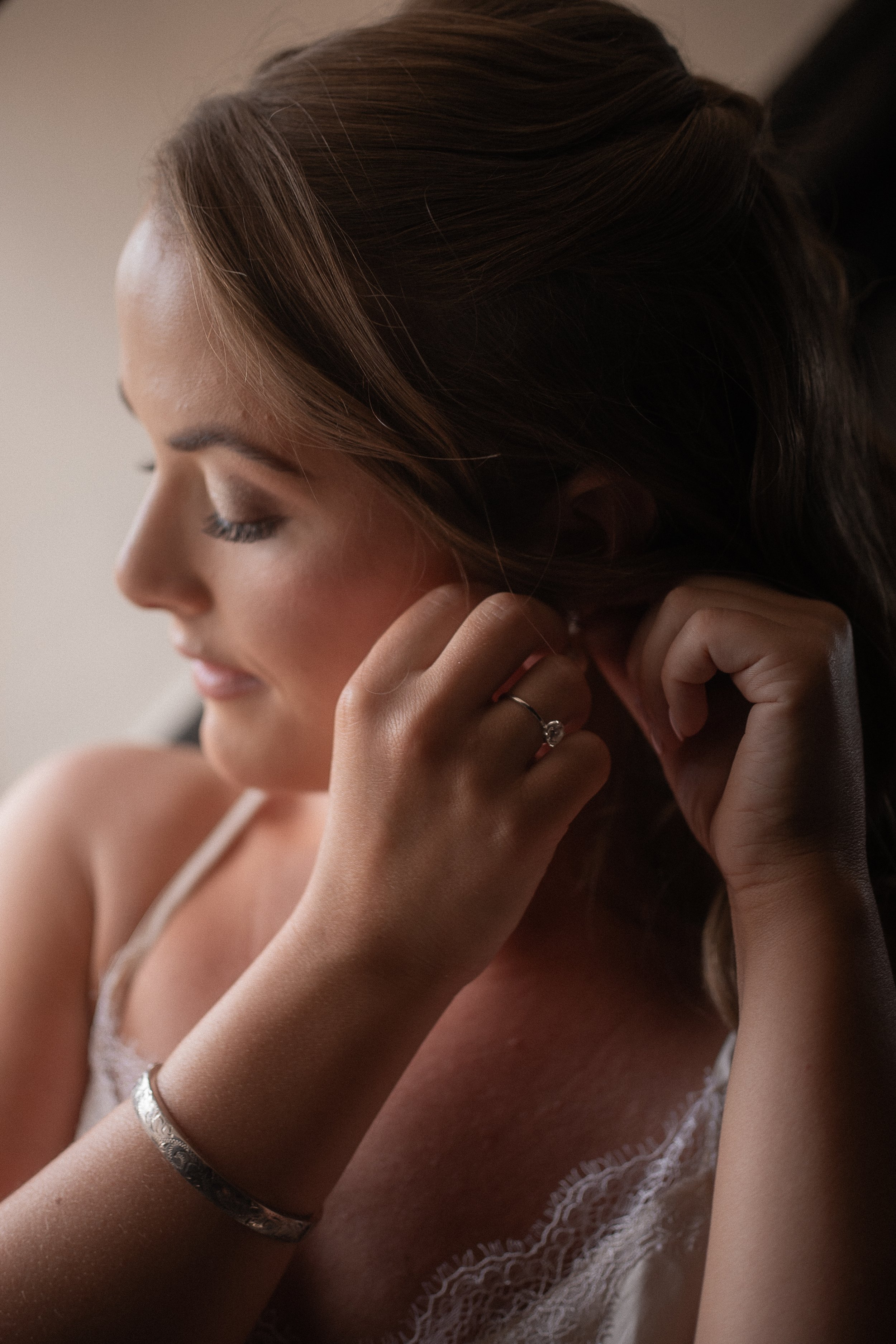 Close-up of a woman with brown hair, closed eyes, wearing a white lace top, jewelry, and resting her hand near her face.