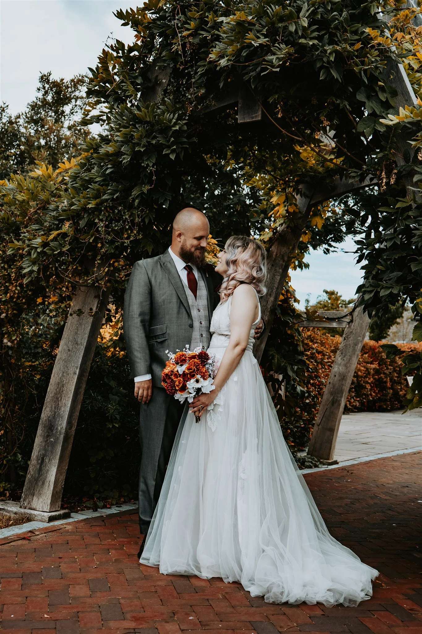 A bride and groom standing close under a wooden arch covered with green and yellow leaves, looking into each other's eyes. The bride is holding a bouquet of orange, burgundy, and white flowers, wearing a white wedding gown, and the groom is wearing a