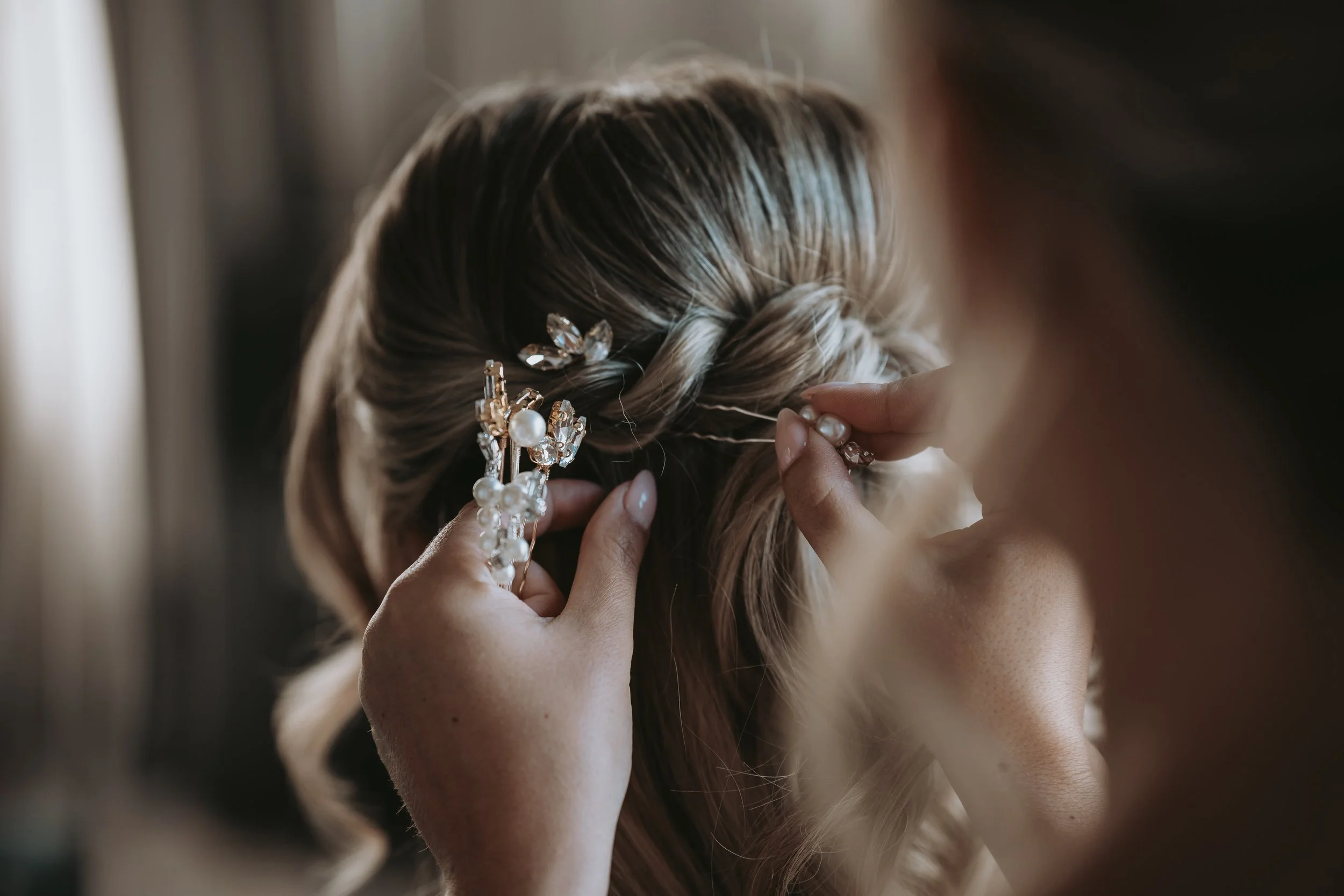 A woman is pinning an intricate pearl and crystal hair accessory into a woman's blonde hair styled in a braid.