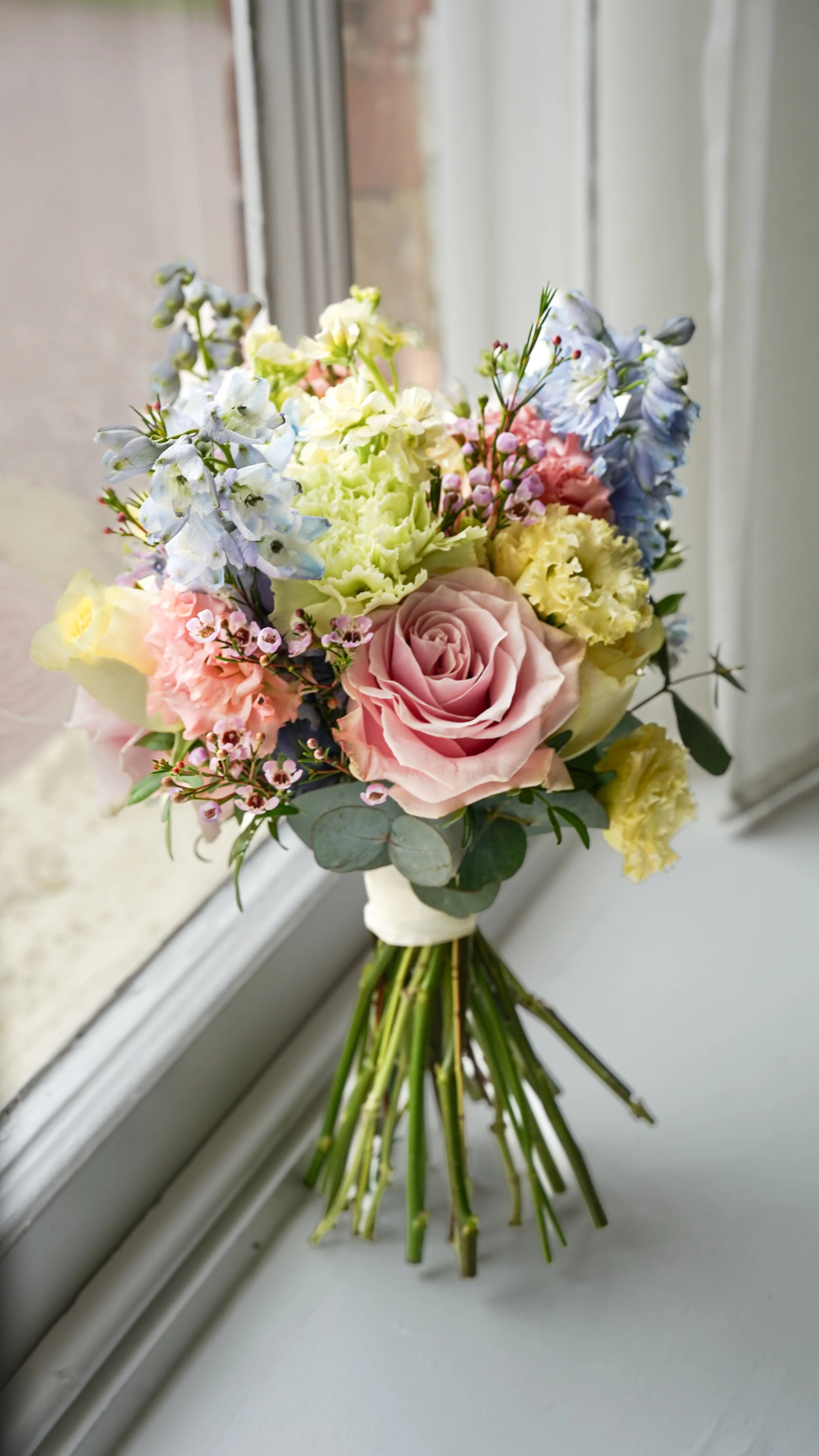 A colorful bouquet of mixed flowers, including pink roses, yellow and white blooms, and blue flowers, arranged on a windowsill.