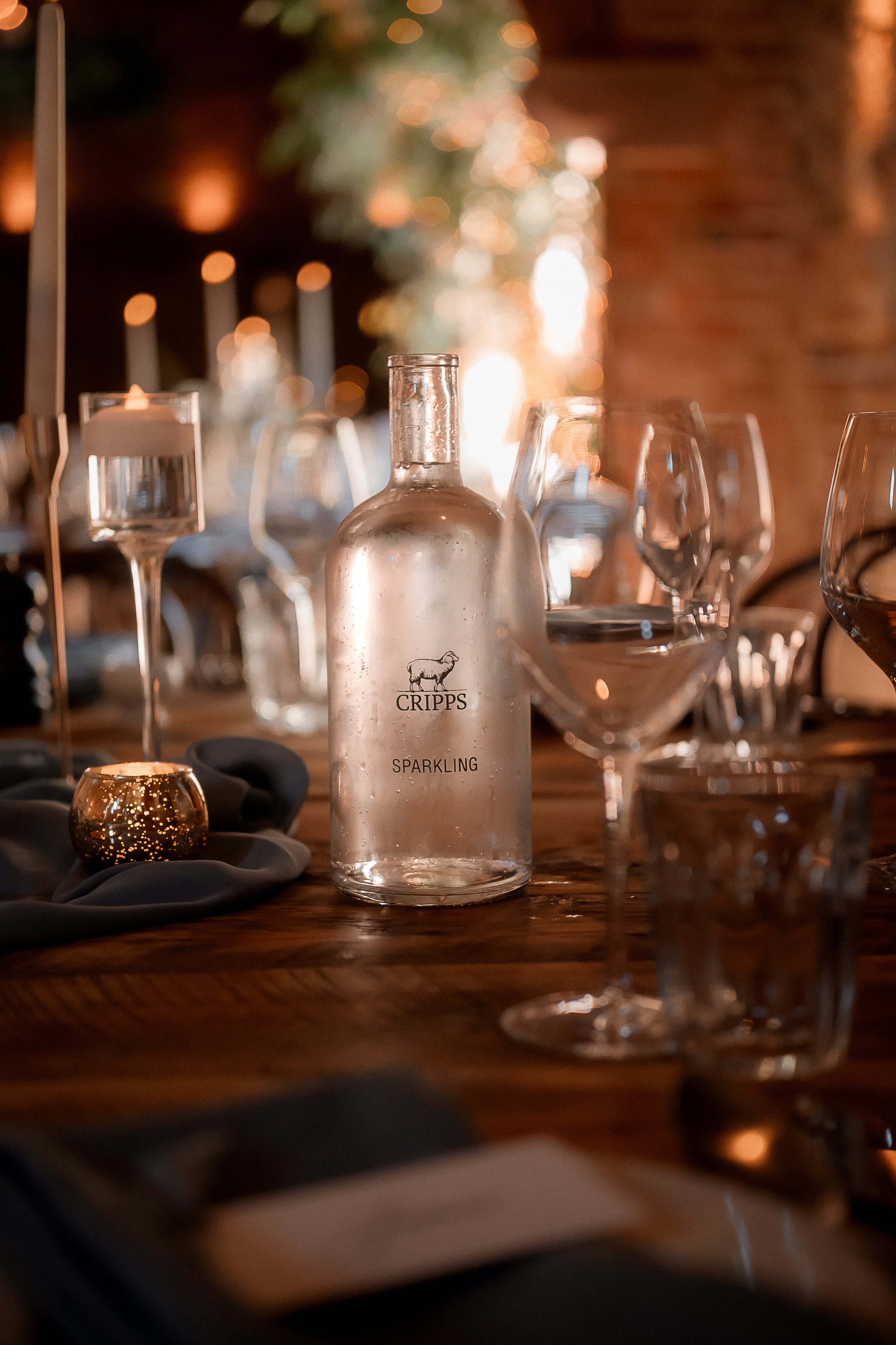 A clear glass bottle of Cripps Sparkling wine on a wooden table, surrounded by wine glasses, candles, and soft lighting in an elegant setting.