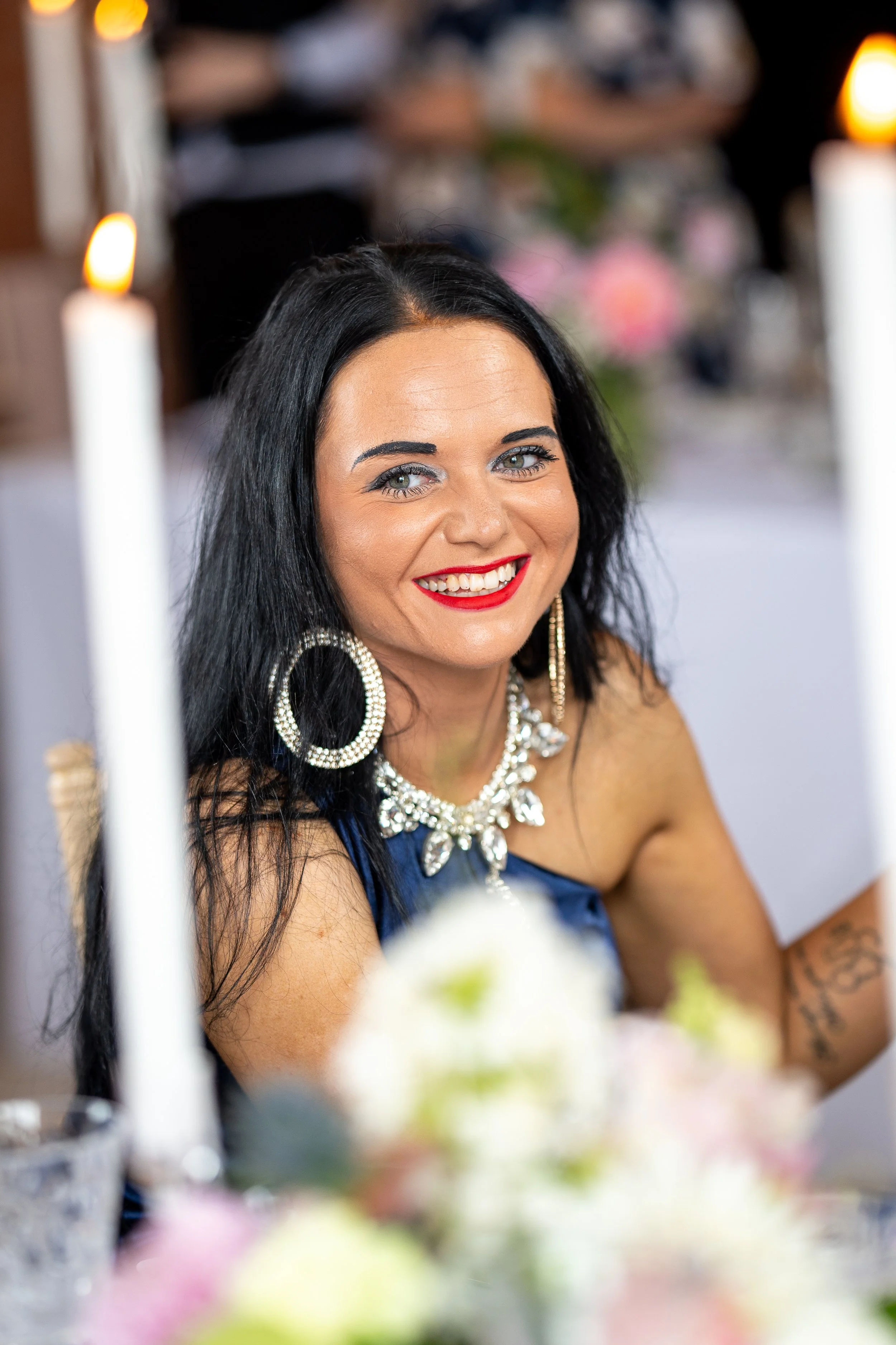A woman with long black hair, bright blue eyes, and red lipstick, smiling at a formal event with candles and floral decorations in the background, wearing large hoop earrings and a pearl necklace.