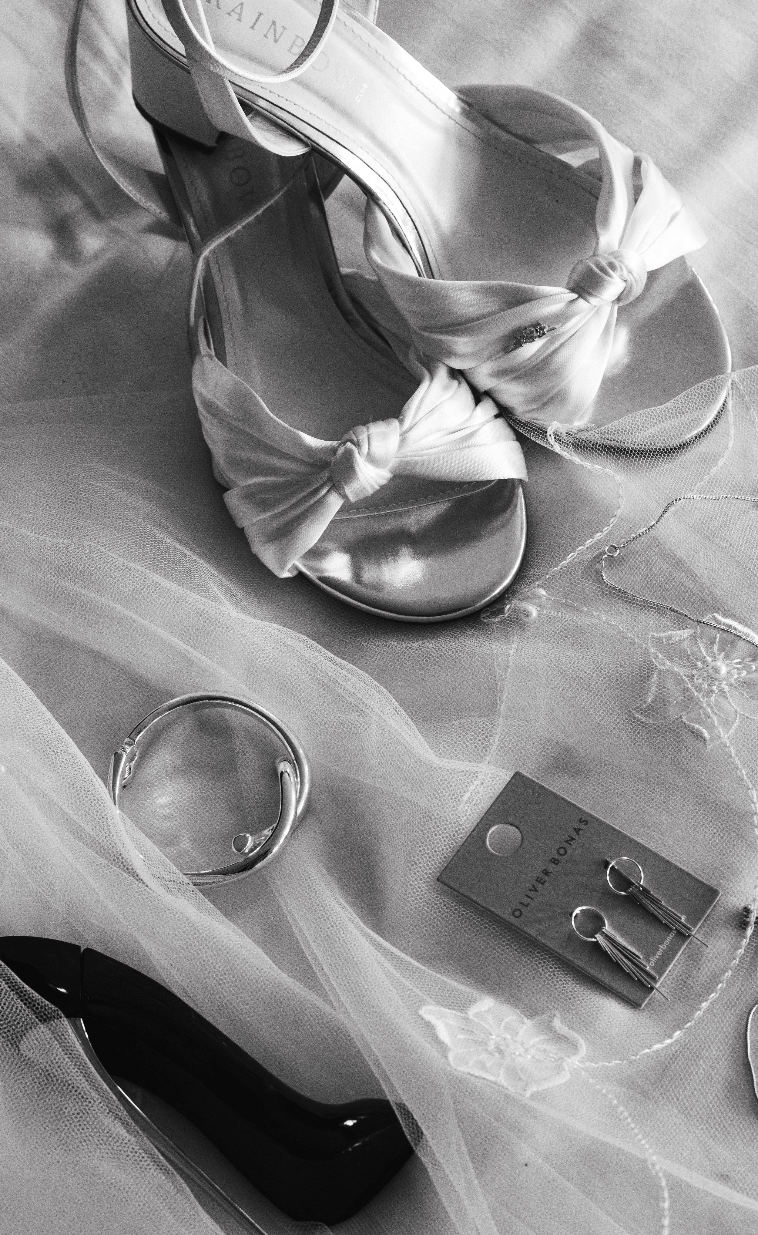 Black and white photo of knotted open-toe bridal shoes, metallic earrings, ring, and a lipstick on sheer fabric.