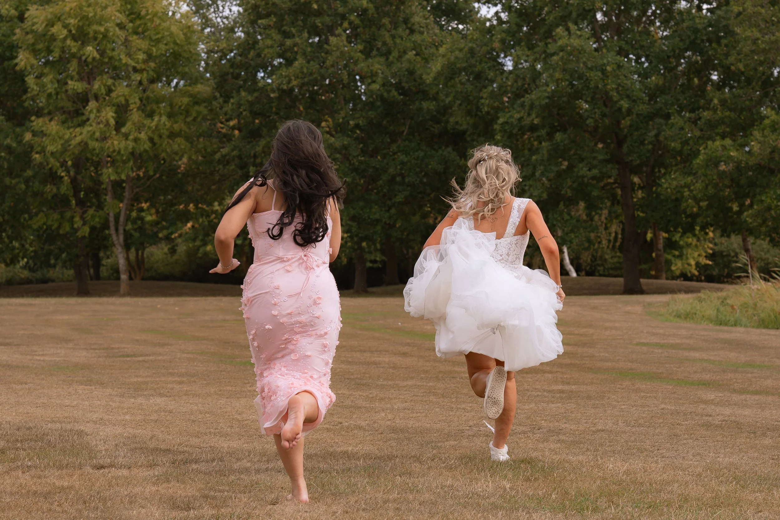 Two women in dresses running across a grassy field, with trees in the background.
