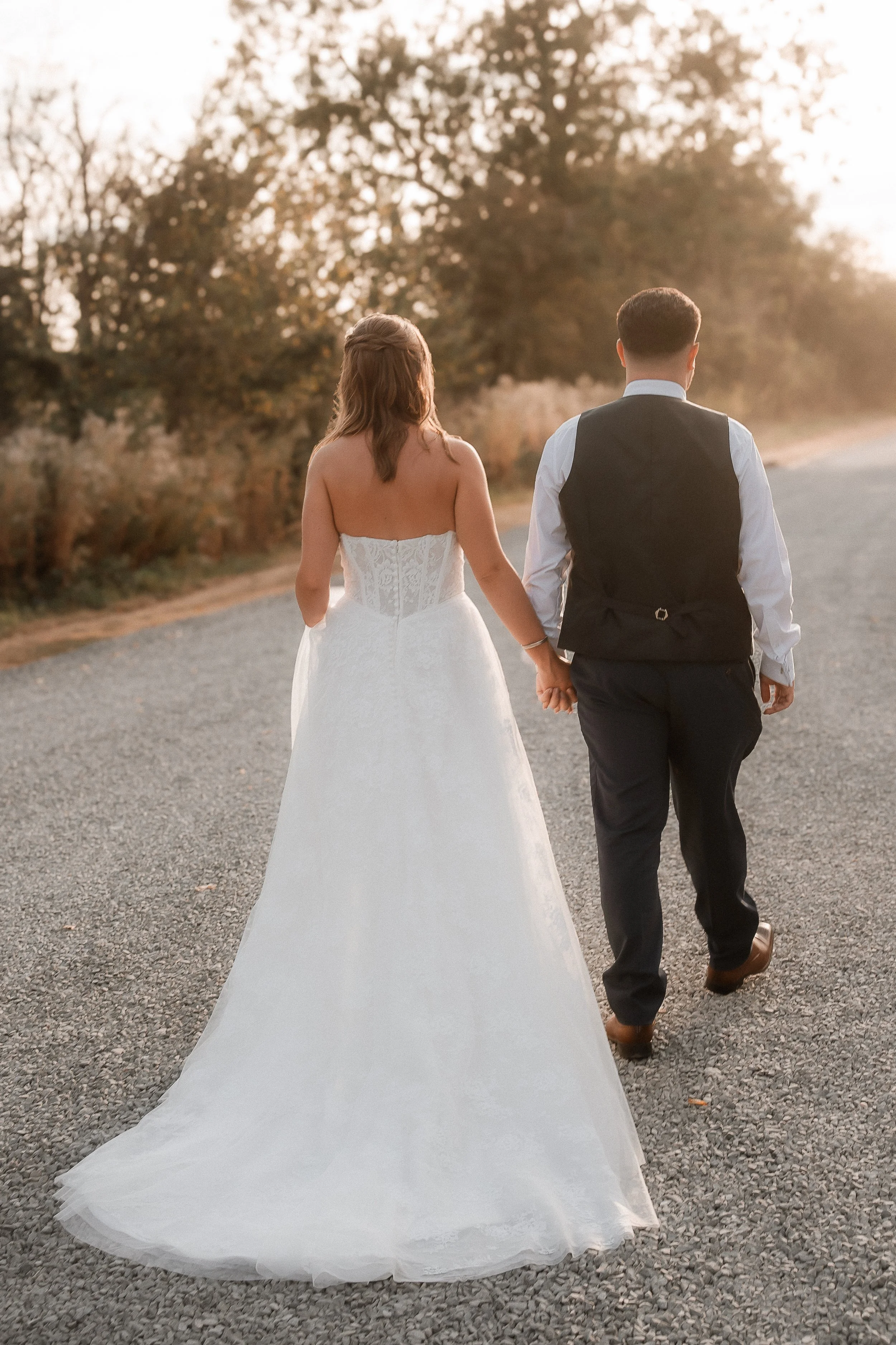 A couple dressed in wedding attire walking hand-in-hand on a gravel road, with trees and sunlight in the background.