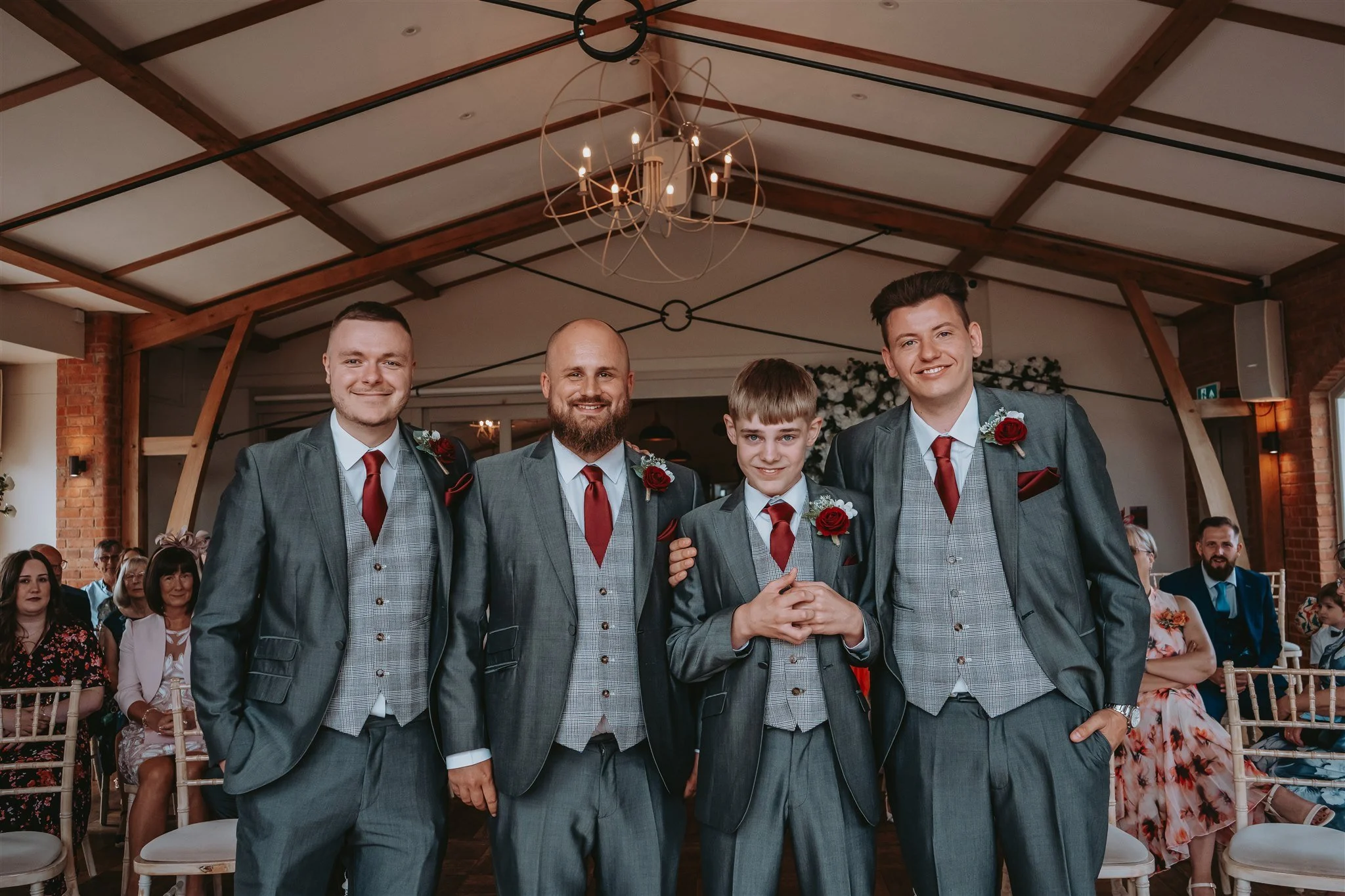 Four men, including a young boy, dressed in gray suits with red ties and red boutonnières, are standing together at a wedding ceremony inside a venue with a wooden ceiling and chandeliers. Guests are seated behind them, some watching the event.