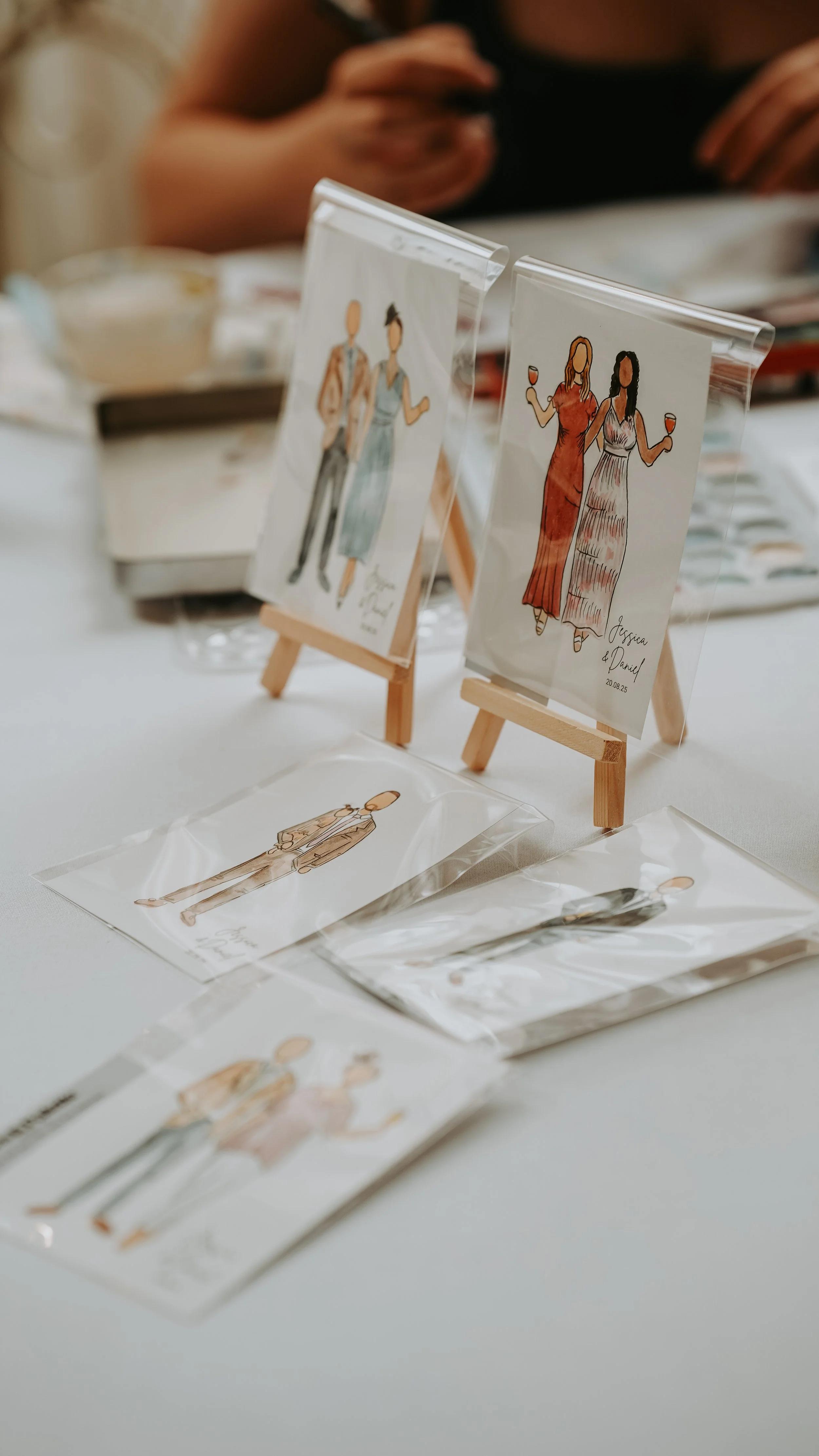 Drawings of wedding attire and couples, some in plastic sleeves, displayed on small wooden easels and paper on a table, with a person in black in the background.
