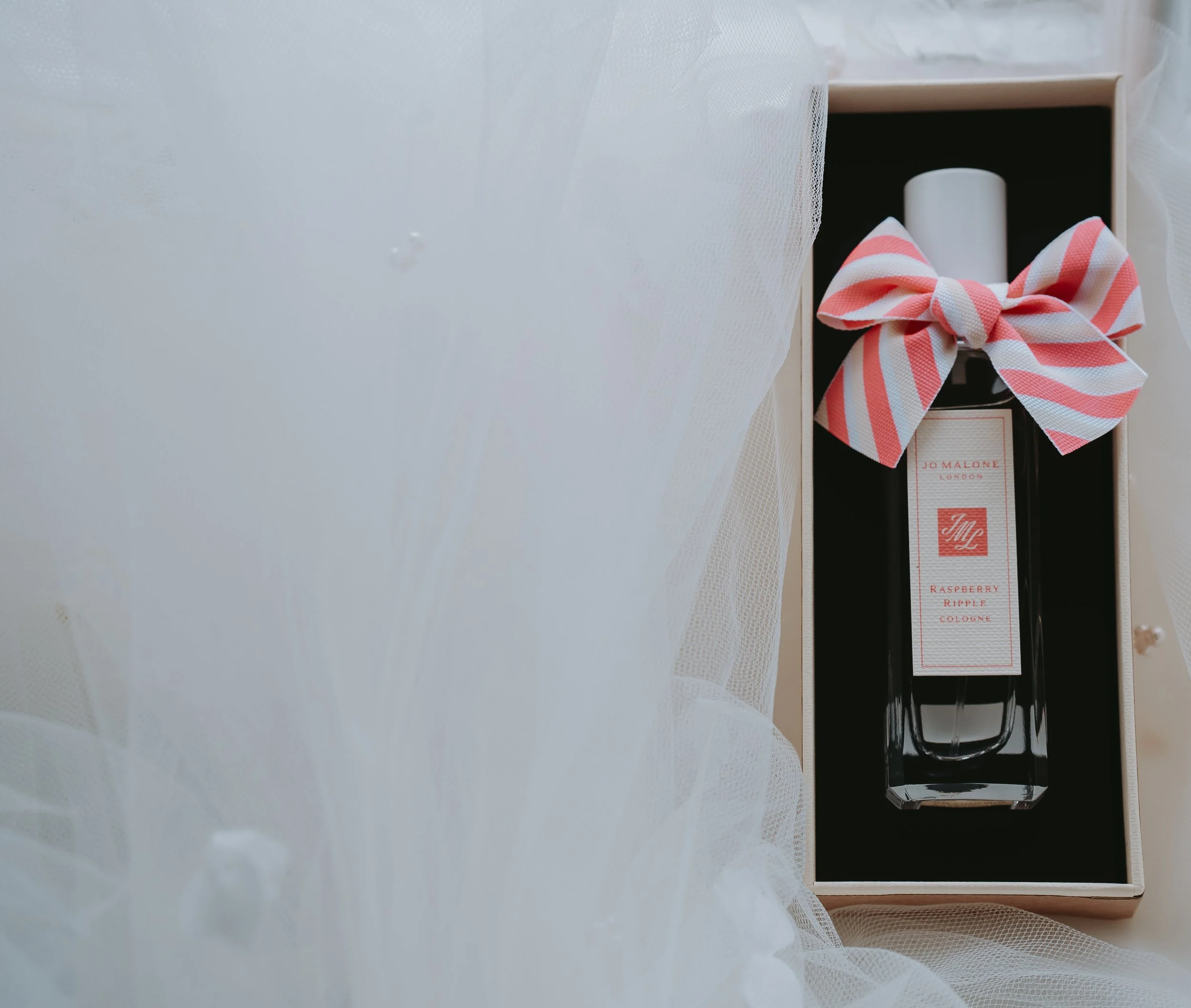 A bottle of Jo Malone Raspberry Ripple Cologne with a pink and white striped bow in a gift box, partially covered by white tulle fabric.