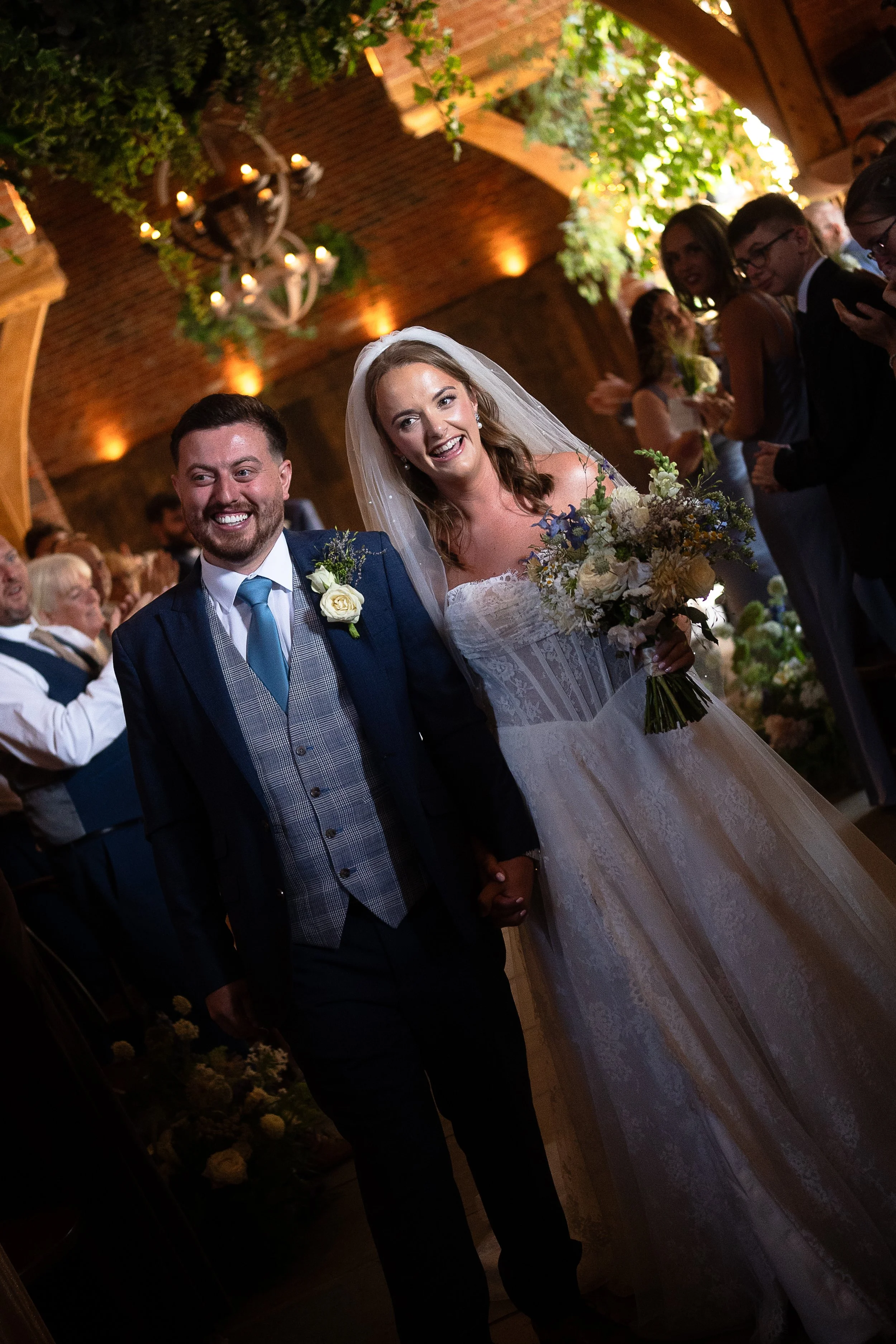 A bride and groom smiling at each other during their wedding reception inside a decorated rustic venue with lit chandeliers and guests in the background.