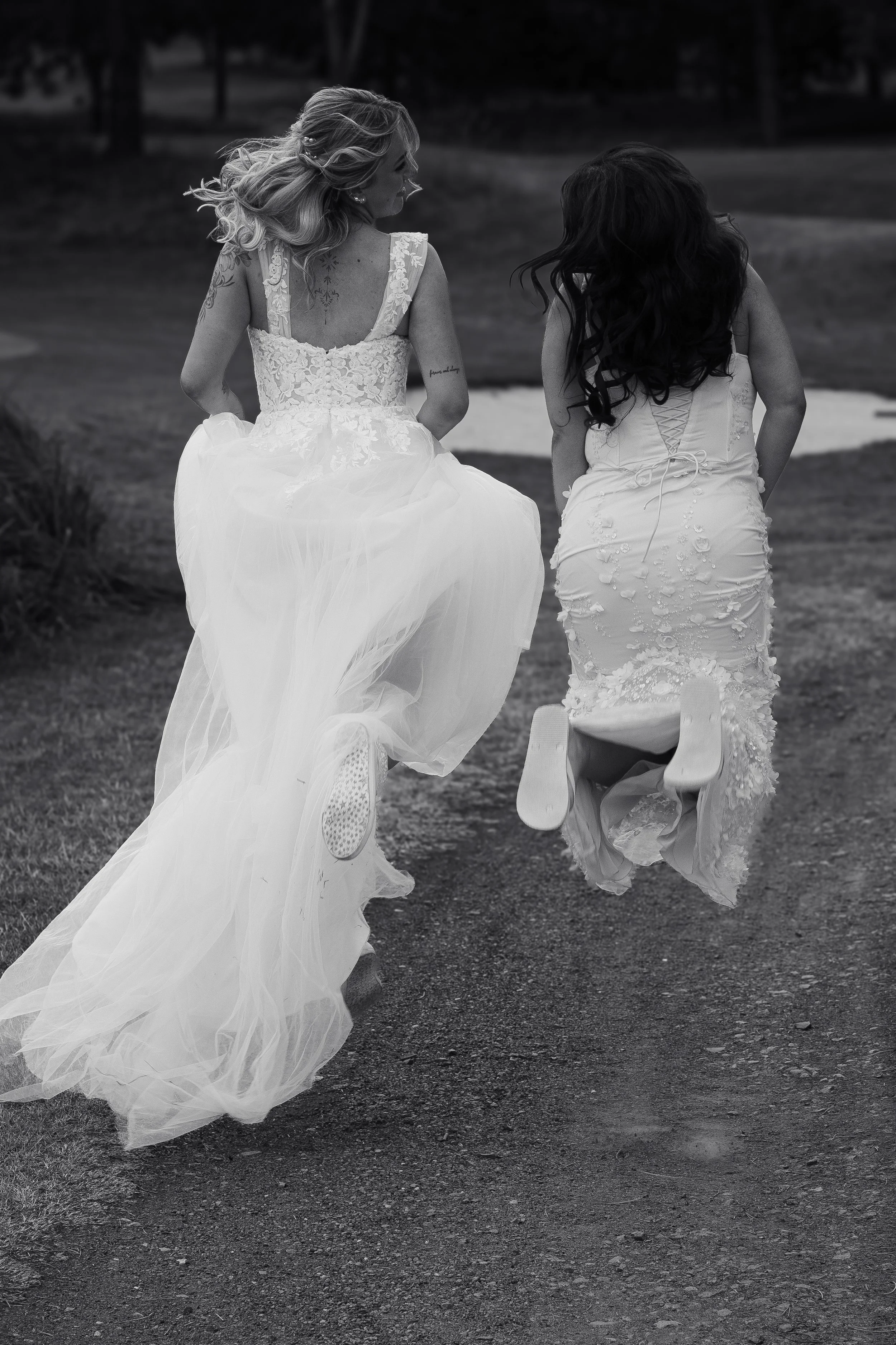 Two women in wedding dresses running on a dirt path, holding hands, with trees in the background.