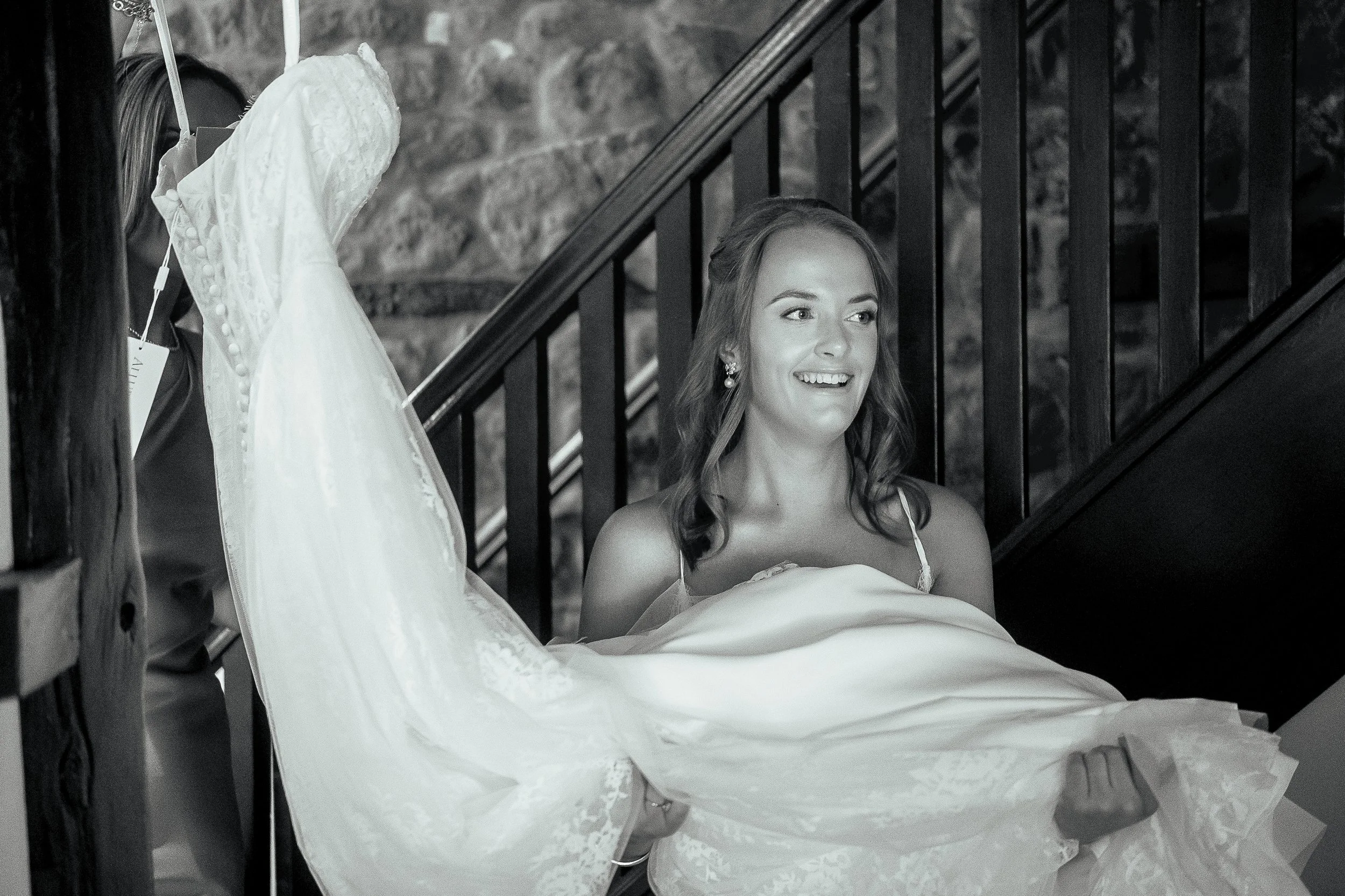 A woman in a strapless dress sitting on the stairs, smiling and looking to her right, with a wedding dress laid out in front of her.