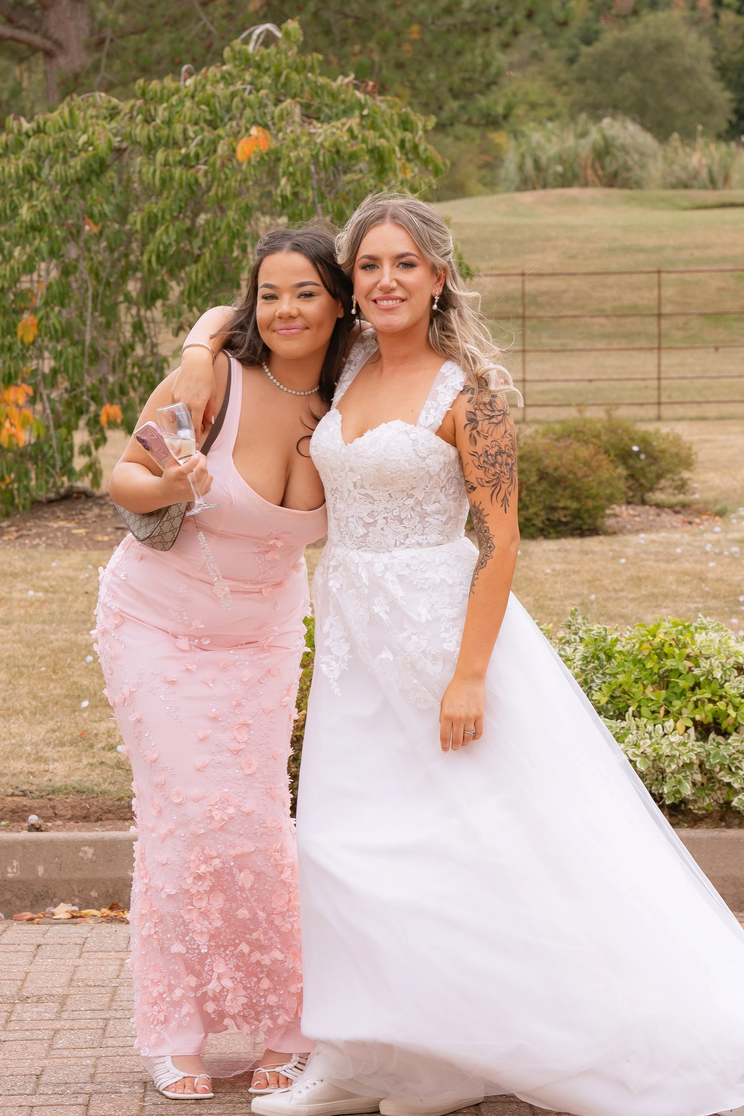 Two women, one in a pink dress and the other in a white wedding gown, standing together outdoors smiling and embracing, with trees and bushes in the background.