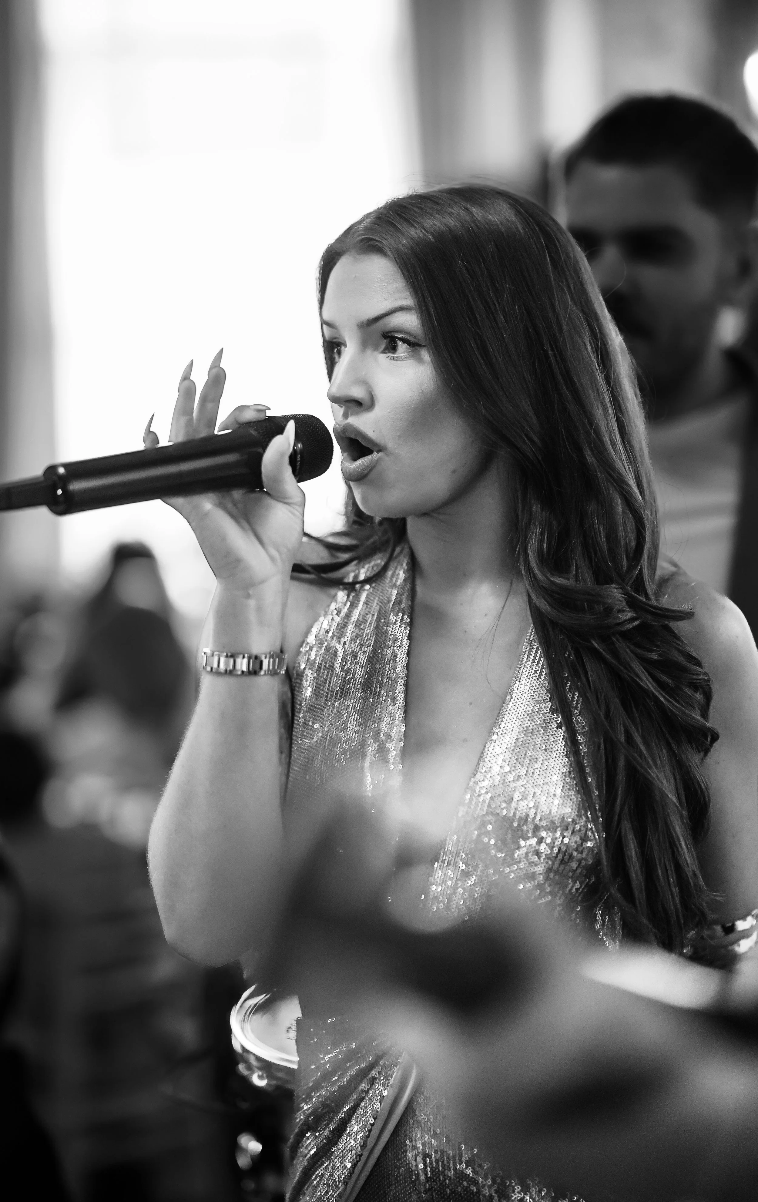 A woman with long wavy hair, wearing a sleeveless, shiny dress, is singing into a microphone at what appears to be a performance or event.