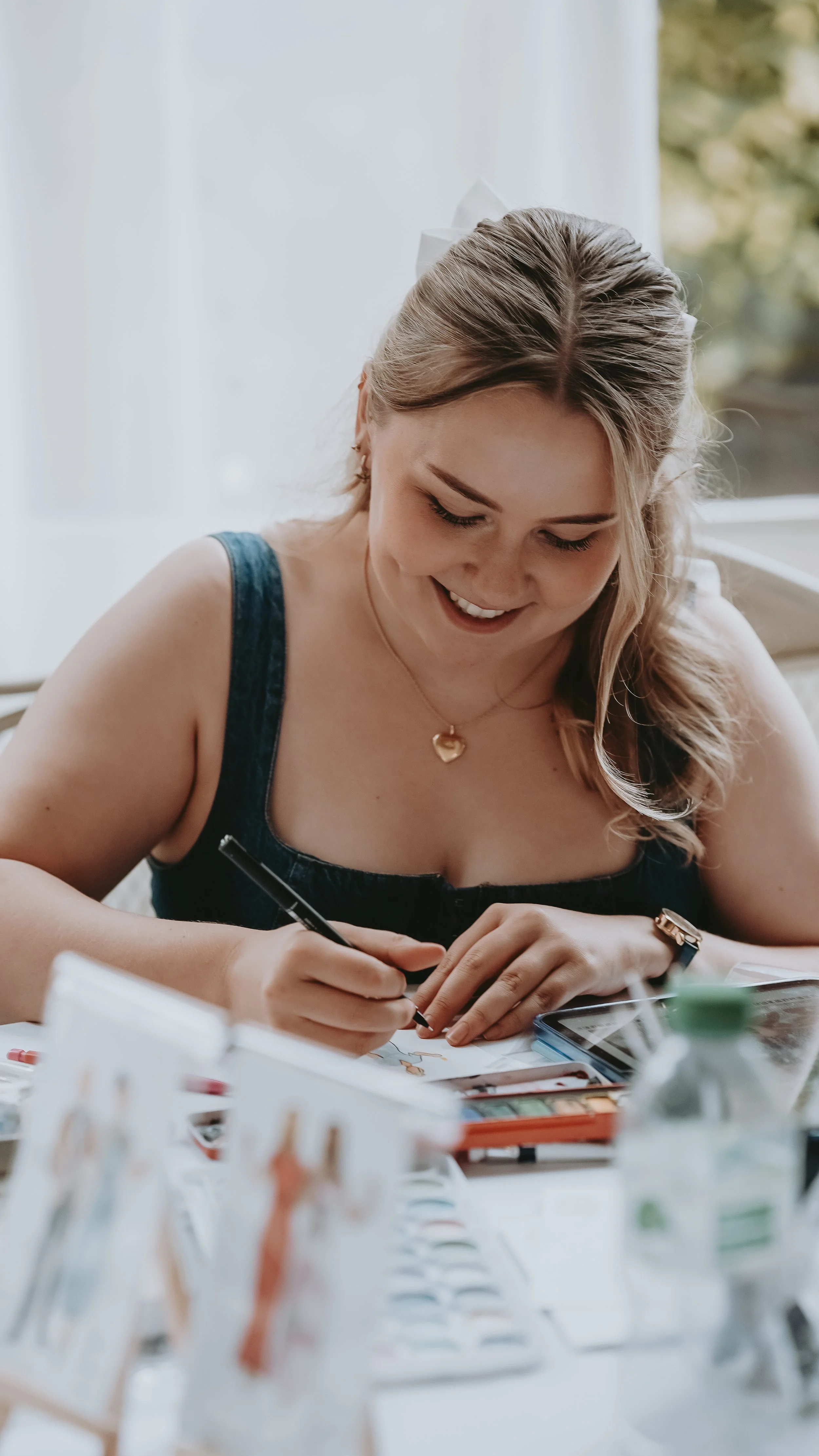 Woman drawing at a desk with art supplies and a smartphone, smiling and looking down at her work.