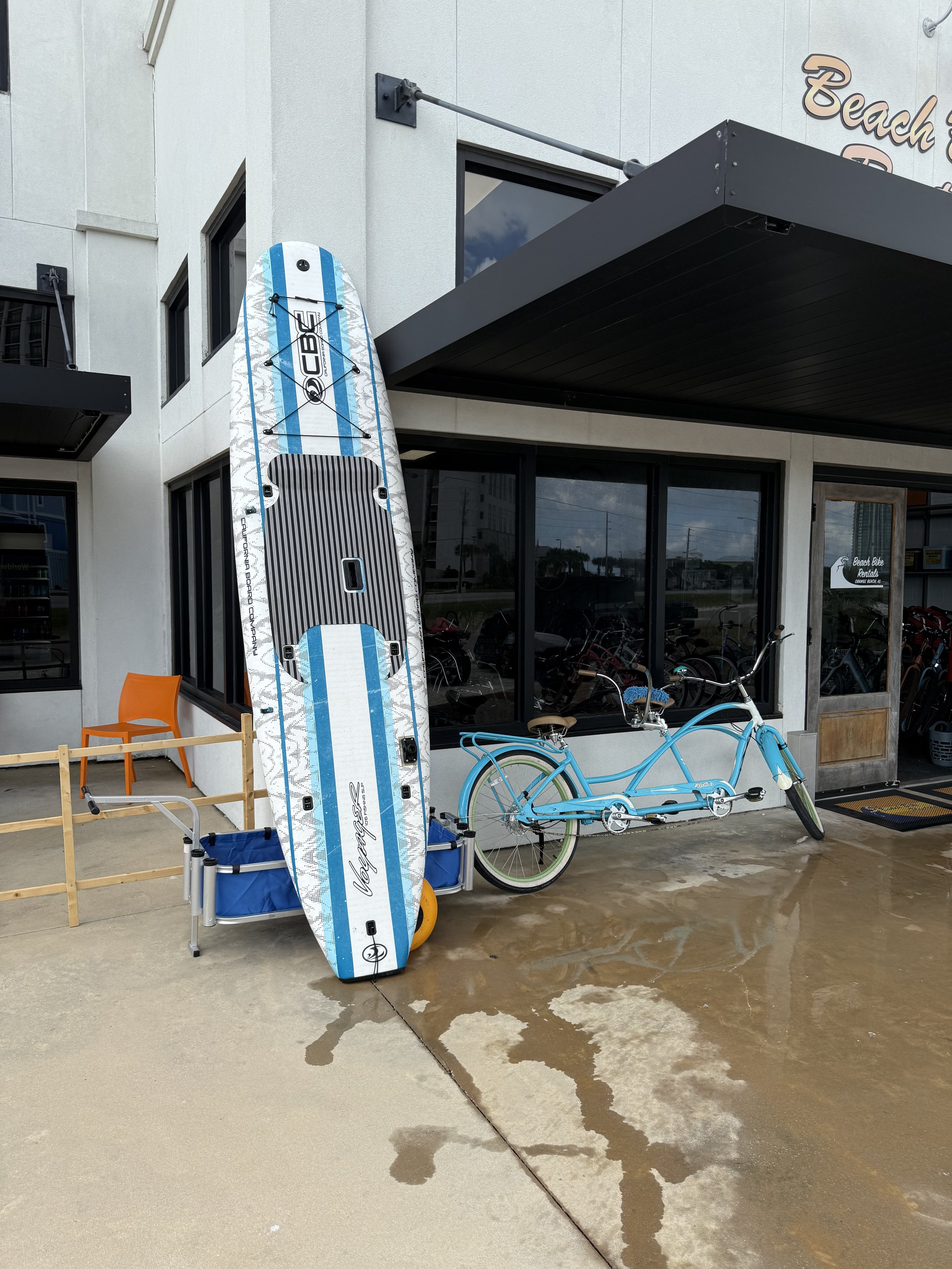 A paddleboard, a bicycle, and a small blue cart are outside a store called 'Beach...' with large windows and a black awning. The paddleboard is propped against the building, the bicycle is parked beside the windows, and the small cart is below the paddleboard.