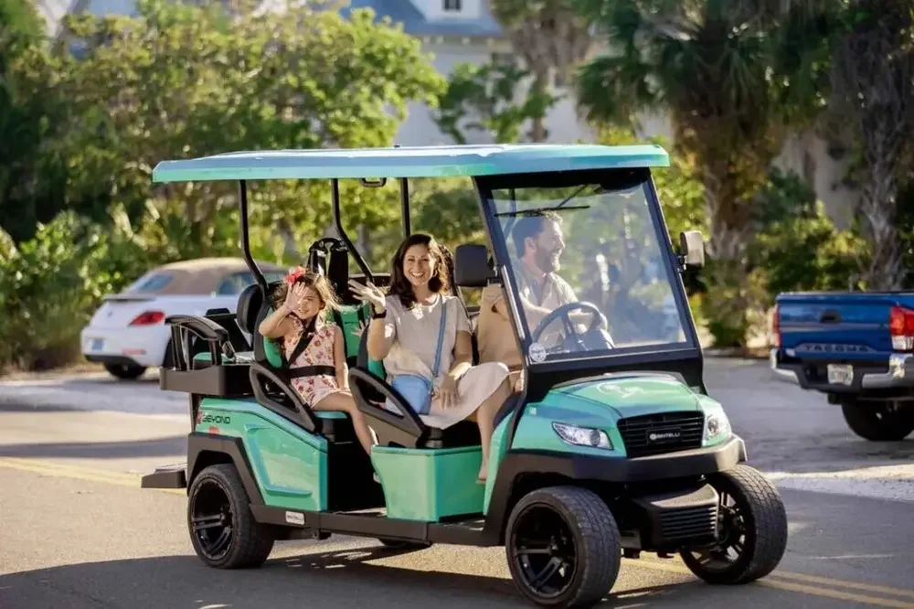 A family of three riding in a teal and black golf cart on a sunny day in a suburban area with trees and parked cars in the background.