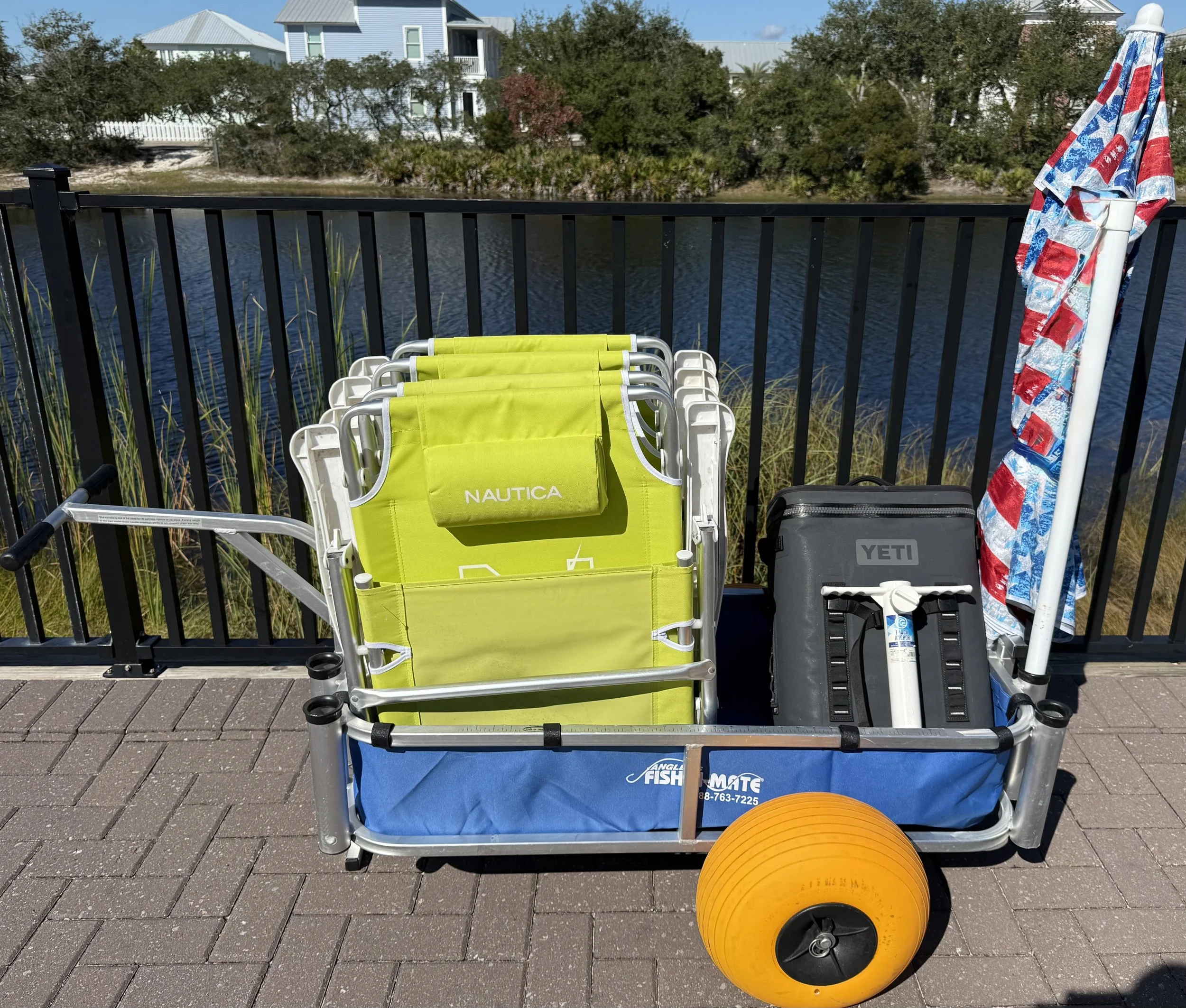 A cart on a sidewalk holding a bright yellow outdoor chair, a black YETI cooler, a folded umbrella with a red, white, and blue pattern, and an orange buoy near a black metal railing overlooking a body of water with houses and trees in the background.