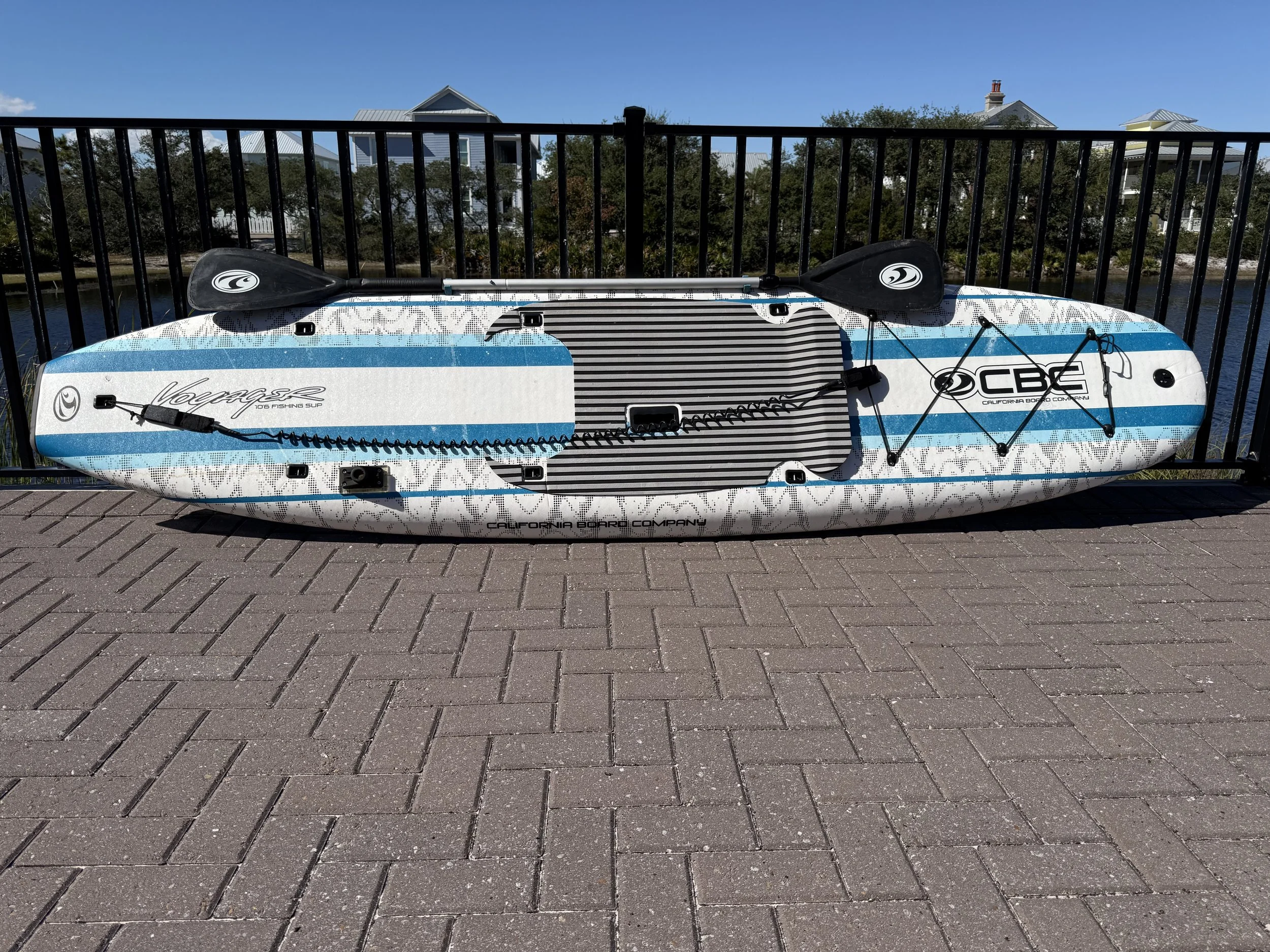 Inflatable paddleboard with two black paddles resting on top, located on a paved surface next to a black railing, with houses and trees in the background under a clear blue sky.