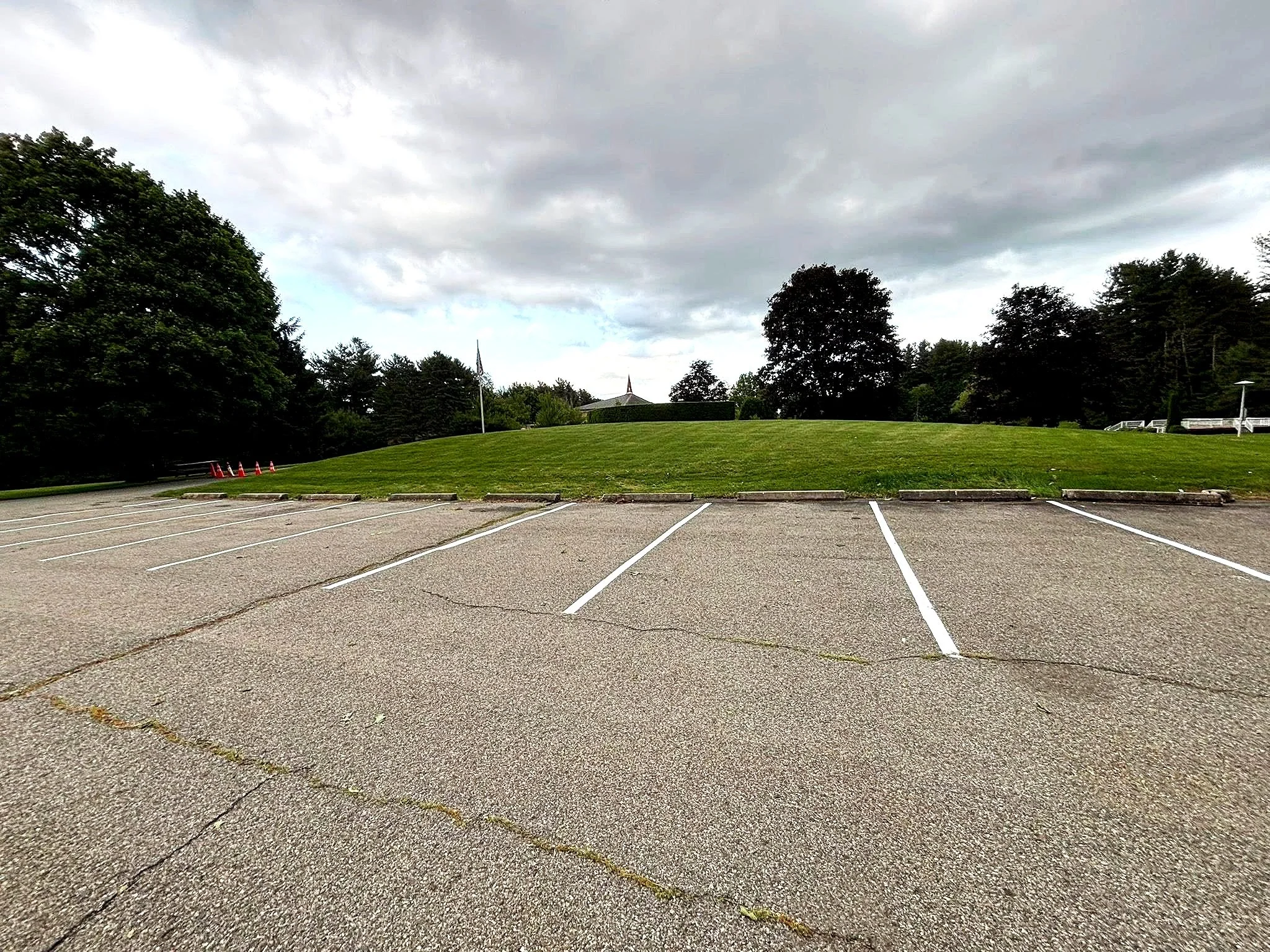 Empty parking lot with a grassy hill, trees, and cloudy sky in the background.