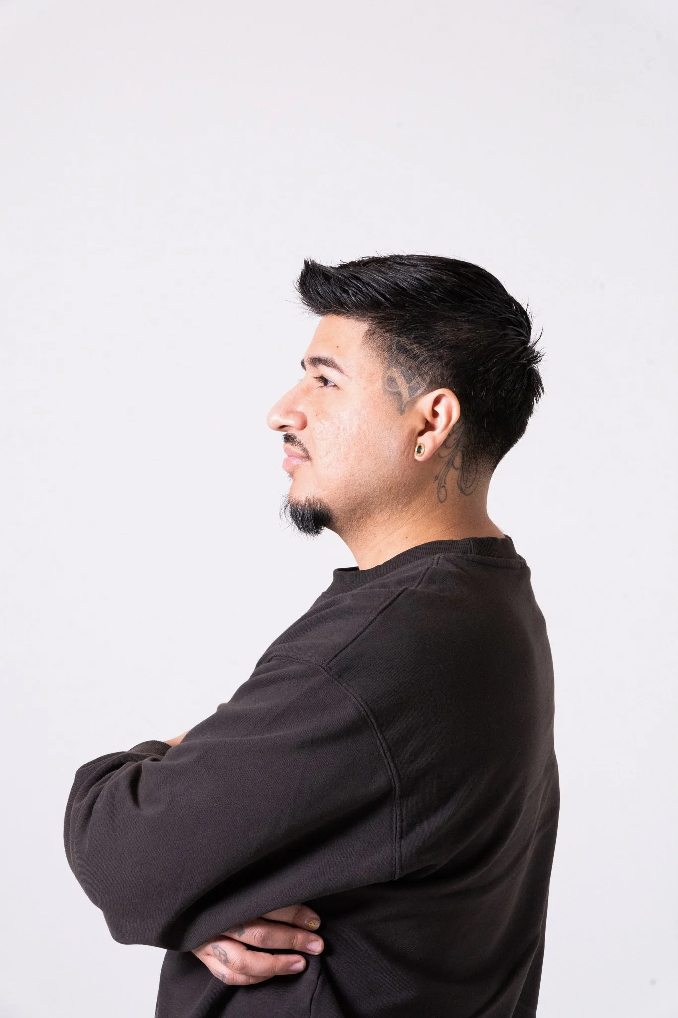 Side profile of a young chicago tattoo artist with black hair, tattoos on his neck and ear, wearing a black long-sleeve shirt, standing against a plain white background.