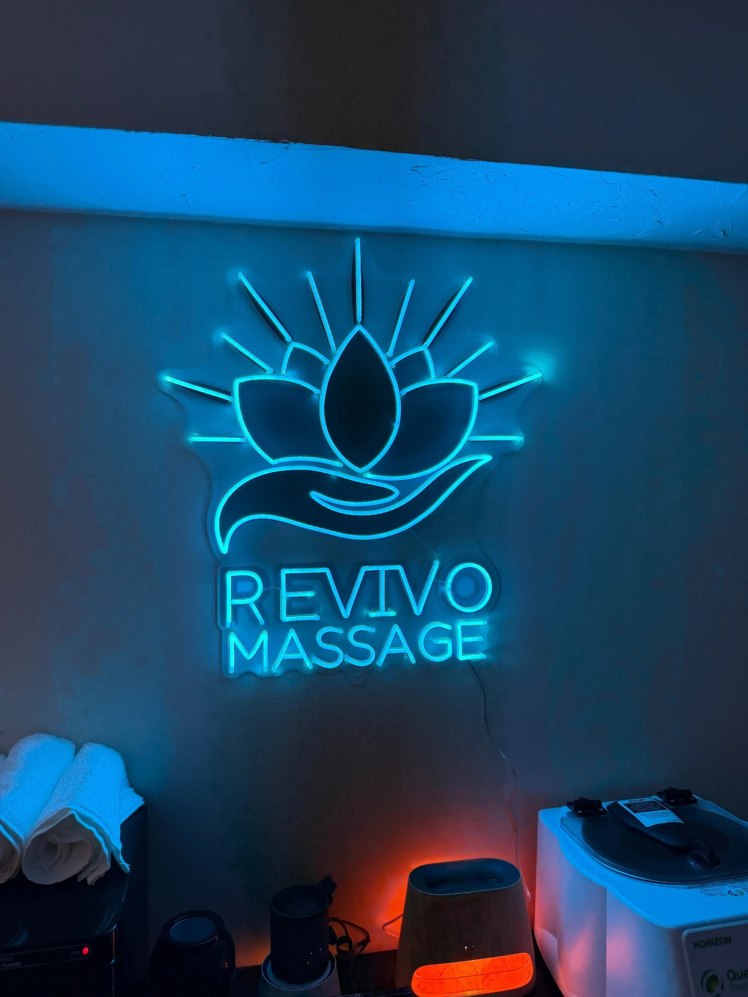 Blue neon sign with a lotus flower and hand logo, reading 'REVIVO MASSAGE', on a wall inside a massage or wellness spa.