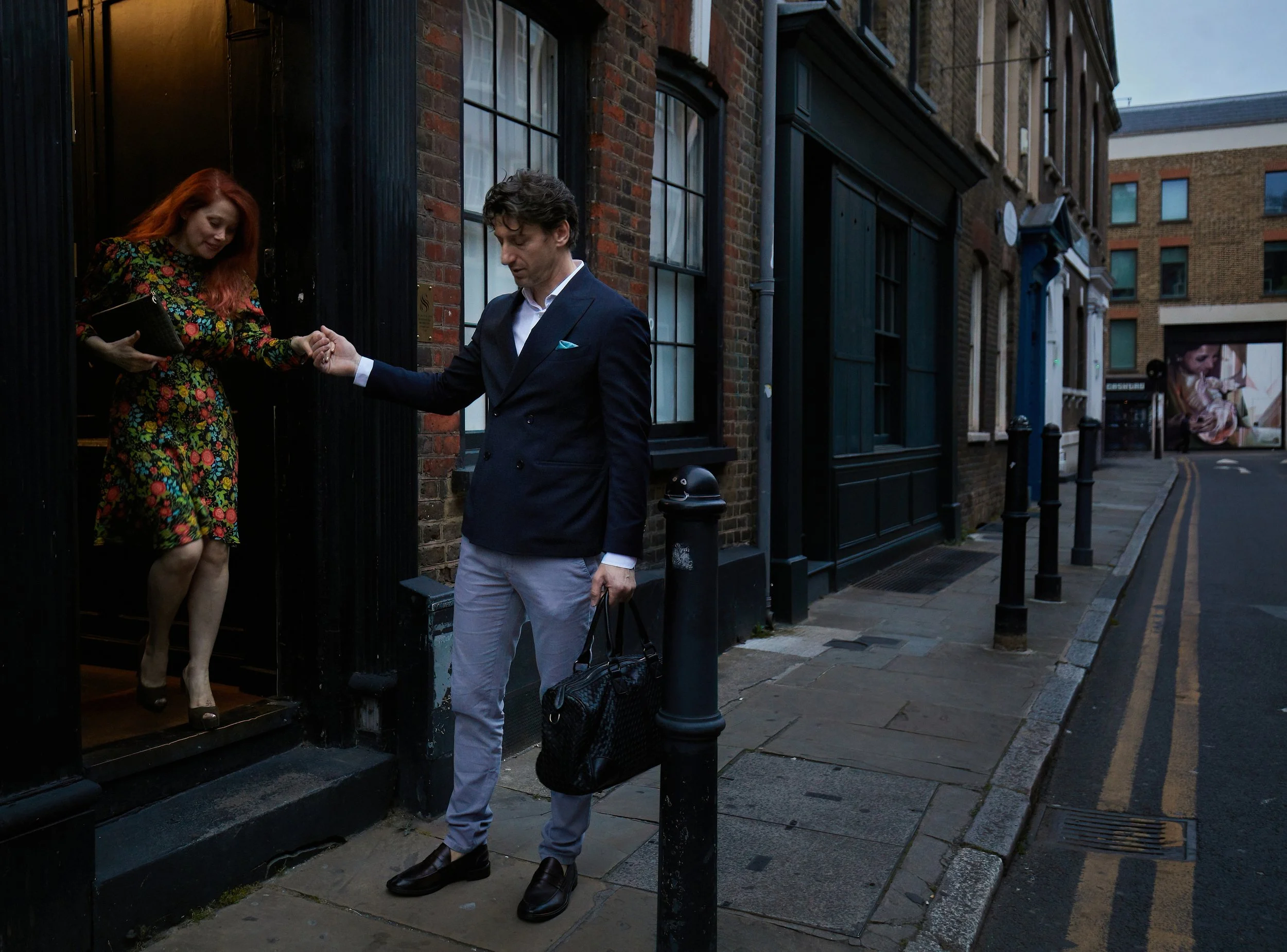 A man in a navy blazer and light gray pants is holding a black bag and holding hands with a woman with red hair wearing a colorful floral dress as she exits a building.