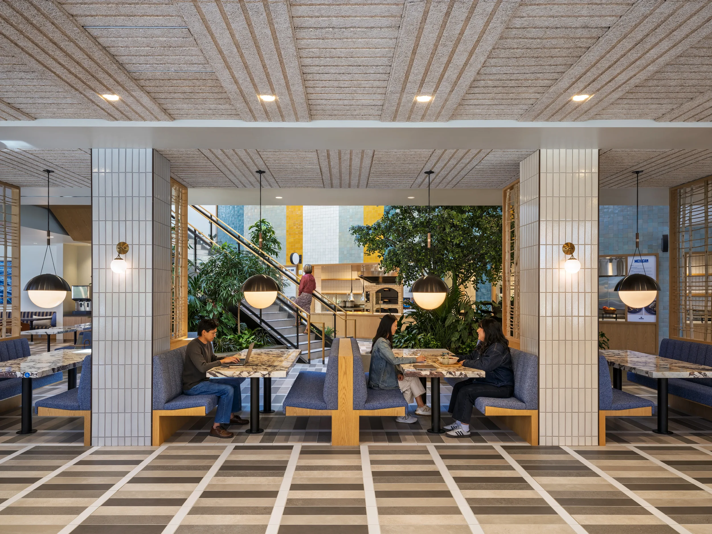 Colorful seating in the Farewinds Food Hall where a man is sitting in a booth working on a laptop and two women sit in the next booth chatting and eating food, with lush plants and a woman walking up a staircase behind them.