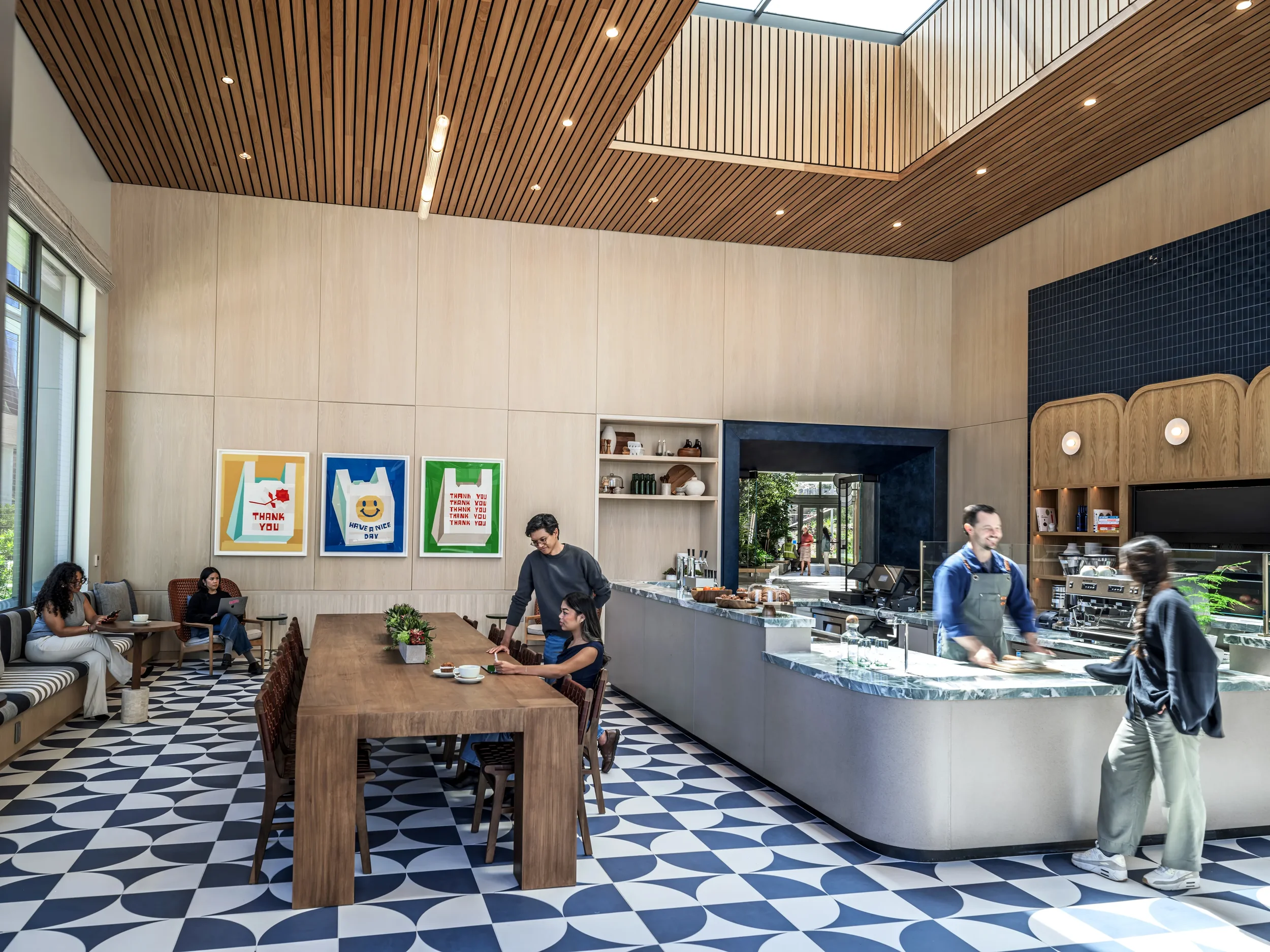Farewinds Market with coffee bar, grab and go drinks and food, patterned tile floor, wooden ceiling, a long wooden table, booths and chairs along walls with round tables, and barista serving a customer.