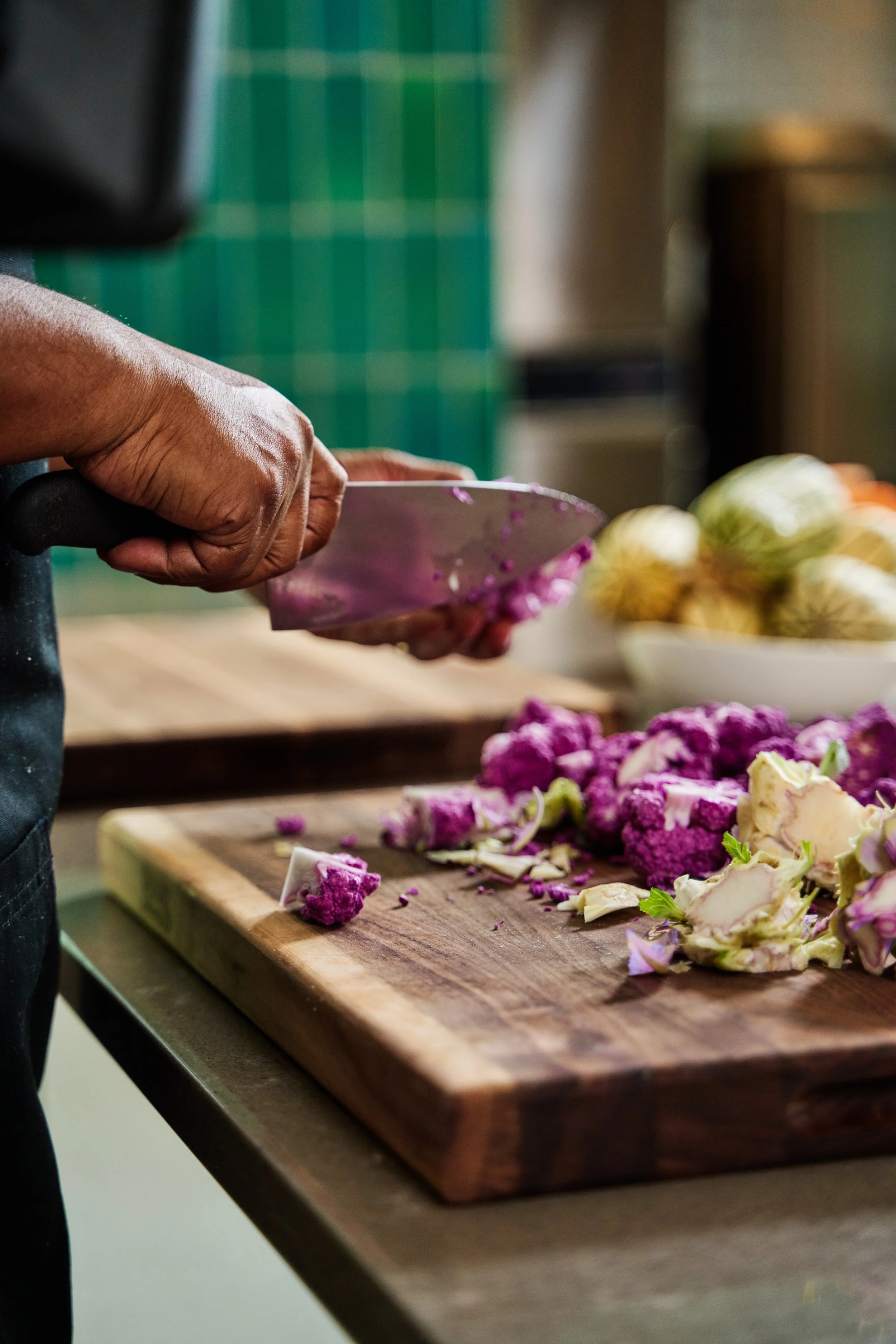  Close up of cutting board and a chef’s hands holding a chef knife and chopping purple cauliflower 