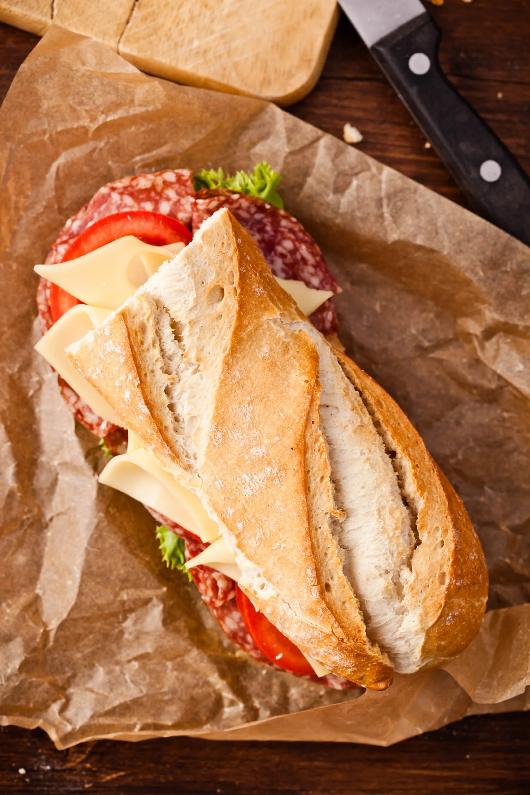  Deli sandwich on baguette with sliced salami, tomato, cheese, and lettuce sitting on brown wax paper 