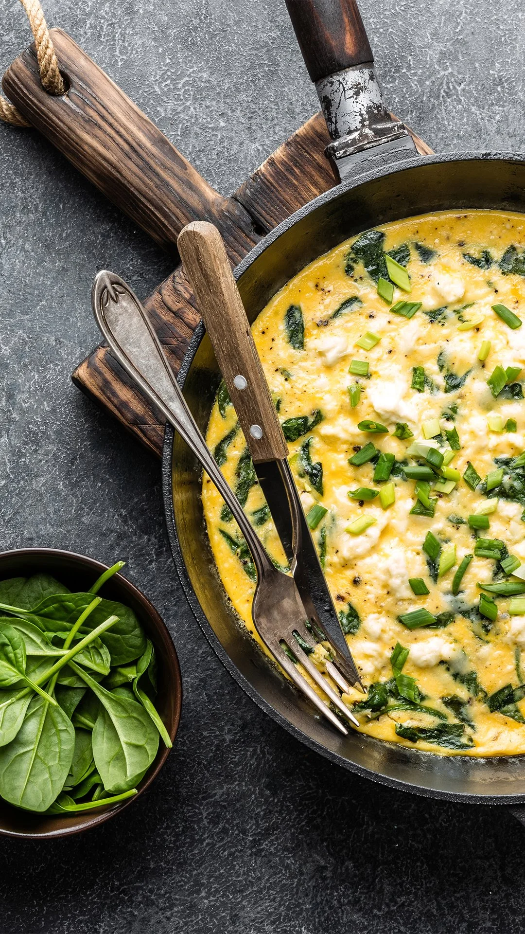  Top view of a fritatta with cheese and herbs in a cast iron skillet with a knife and fork stick into it, next to a small dish of fresh spinach. 