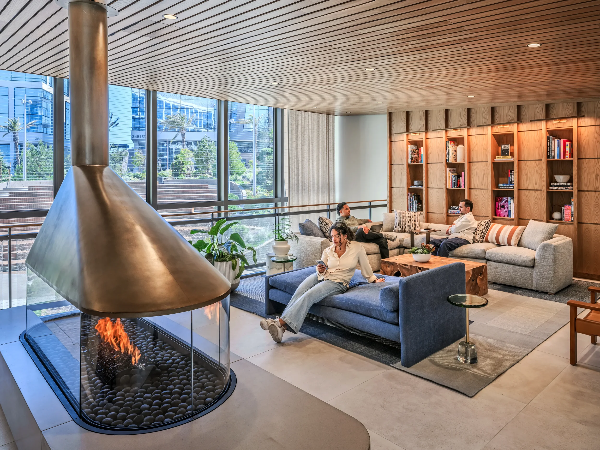 Customers relaxing on couches in front of modern bookshelves in the reservable lounge, with a glass-walled fireplace and hood and window walls looking out onto a landscaped outdoor area.